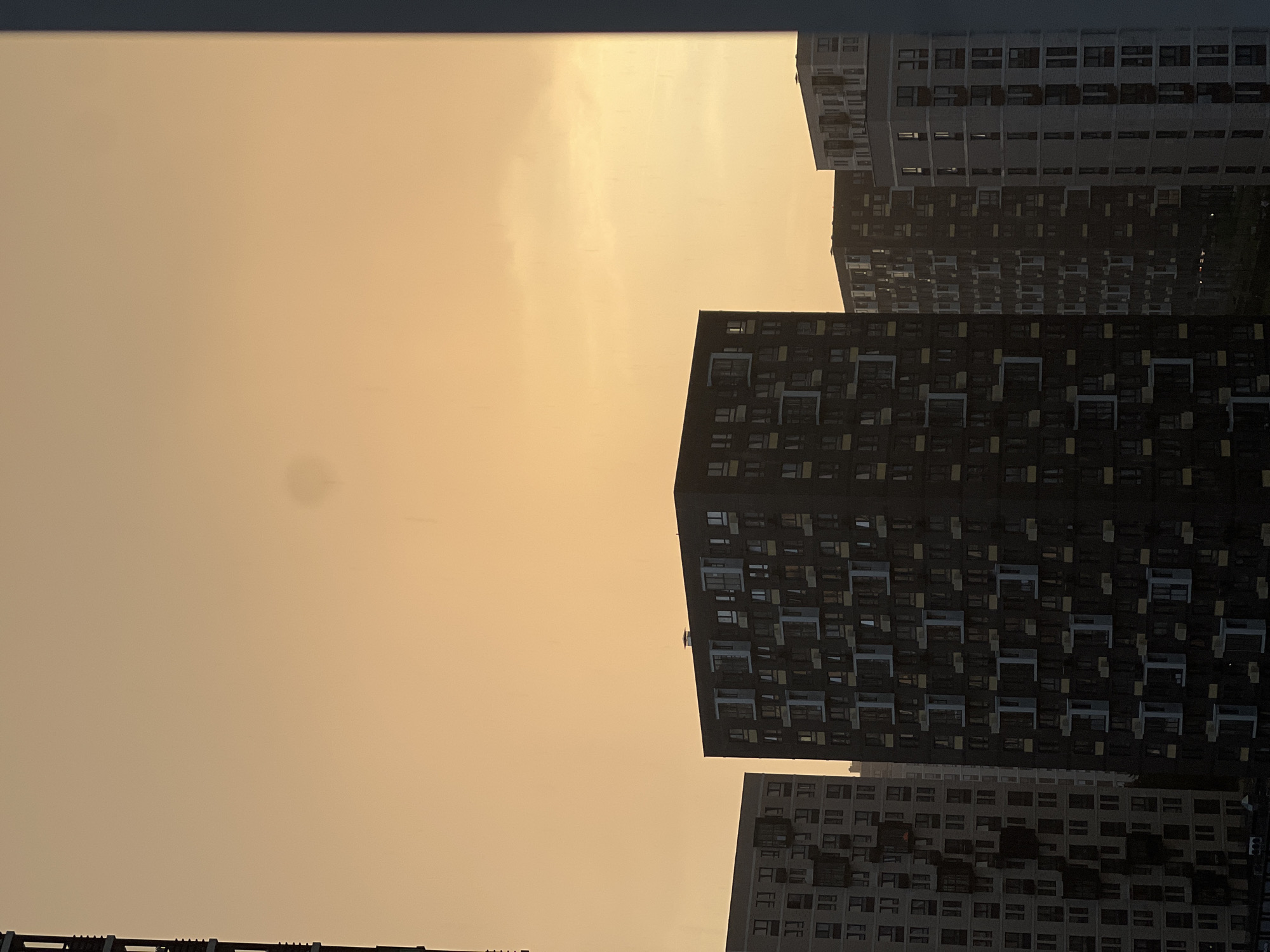 The image depicts the urban landscape of Khimki, Russia, with a focus on high-rise residential buildings silhouetted against a hazy, golden sky, likely indicating a sunrise or sunset. The buildings are characterized by repetitive window patterns and differing shades of gray and black. The scene suggests a dense urban environment. There are no people visible in the image. A partial view of a window frame is visible at the bottom.