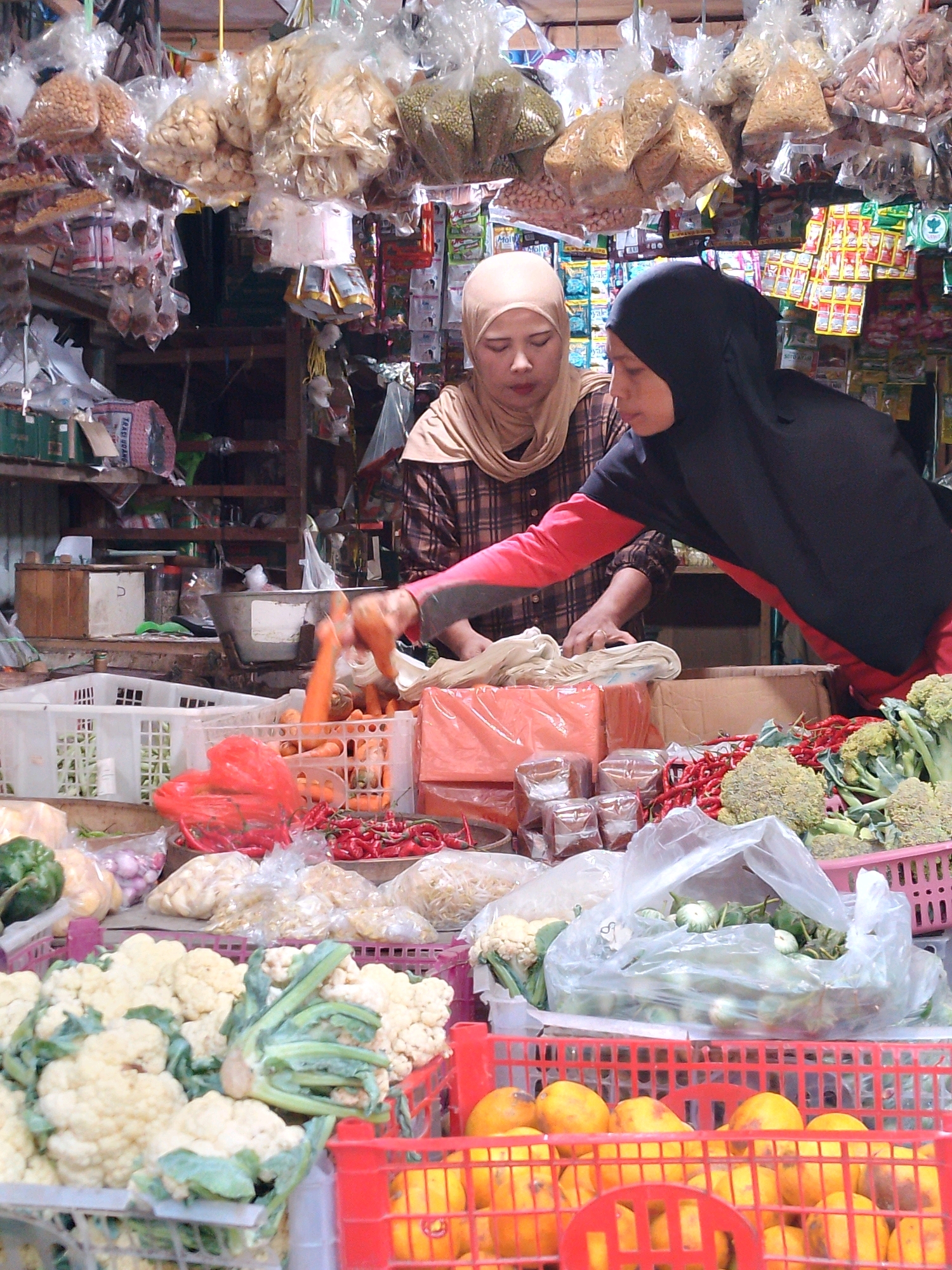 Two women, wearing hijabs, are at a market stall in Kertosono, Indonesia. The woman on the left, wearing a light brown hijab and a checkered shirt, is looking down at the counter. The woman on the right, wearing a black hijab and a red and black garment, is reaching for carrots from a white plastic crate.
The stall is filled with fresh produce and packaged goods. In the foreground, there are baskets of cauliflower, a red crate filled with yellow fruits, and a plastic bag of green vegetables. Behind the women, there are crates of red chilies and broccoli. Packaged snacks and dry goods hang from the ceiling above the stall. The market appears to be an open-air structure with wooden shelves and a metal roof.