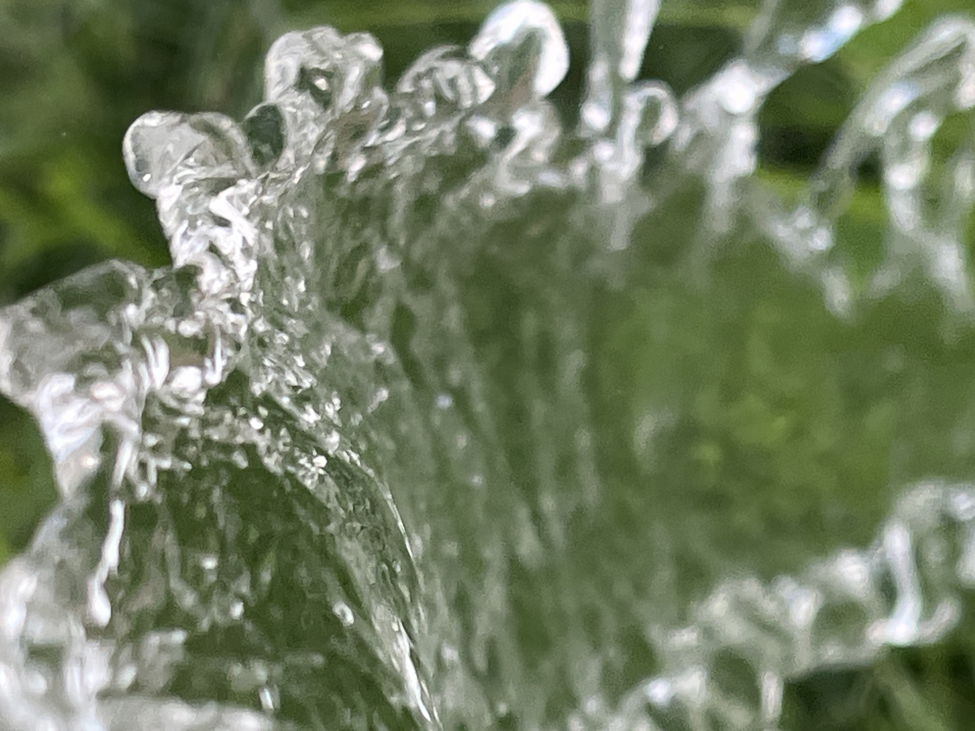 The image shows a close-up of water flowing, with green foliage blurred in the background. The water is in motion, with various shapes and forms, including droplets and streams, visible. The lighting suggests an outdoor setting, possibly during the day. There are no people or discernible man-made structures in the image. The location in Krasnyye Barrikady, Russia, is not visually represented in the photograph.