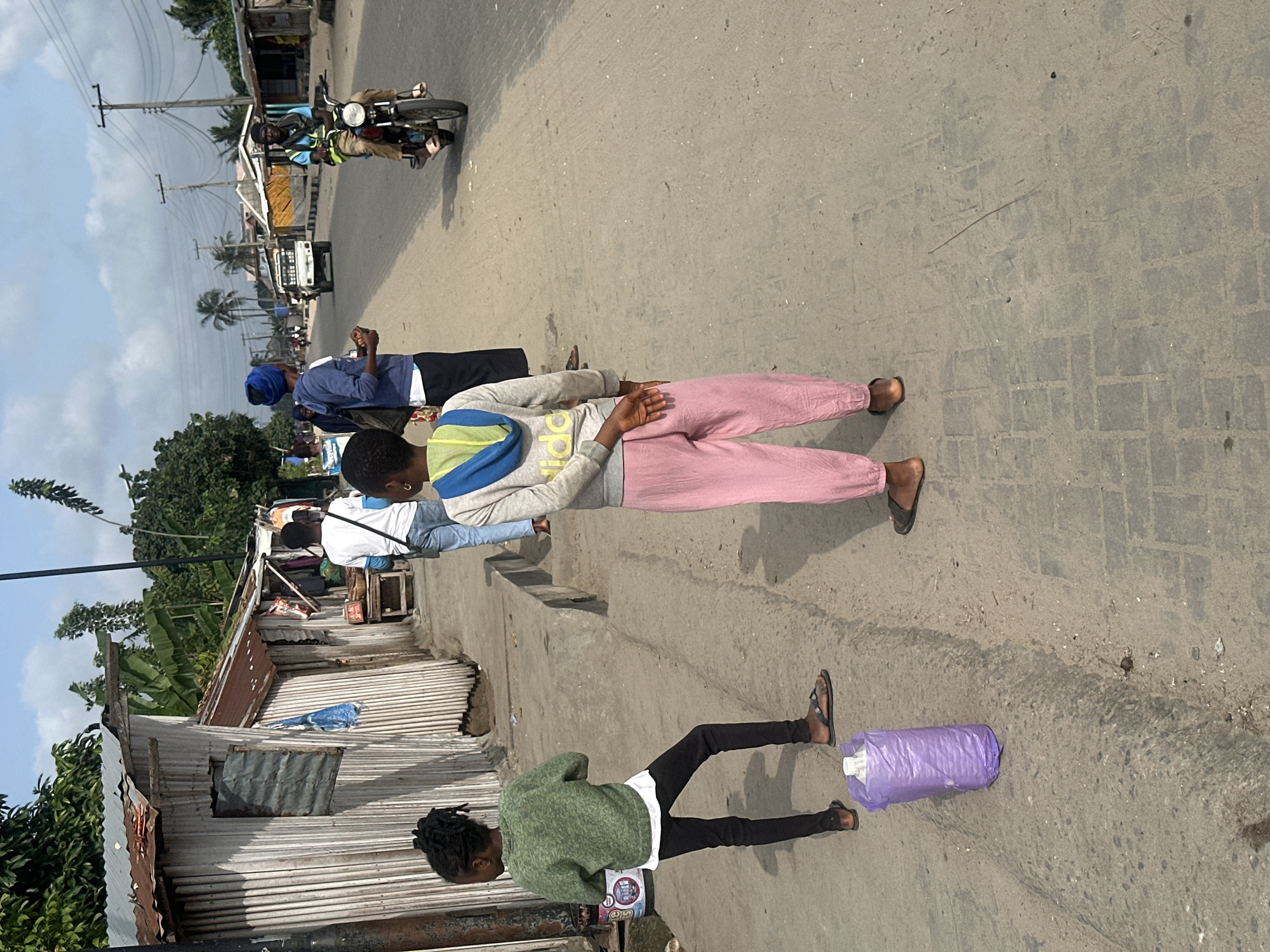 This is a street scene in Badagry, Nigeria. In the foreground, two young girls are standing on a paved sidewalk. The girl on the left wears a green sweater and black pants, and the girl on the right wears a gray hoodie and pink pants. Behind them, a man on a motorcycle wearing a reflective vest rides down the street. Several other people are walking or standing along the sidewalk, and a white truck is visible further down the road. The buildings lining the street appear to be small, informal structures, many with corrugated metal roofs. The overall impression is of a busy, everyday street in a West African city.