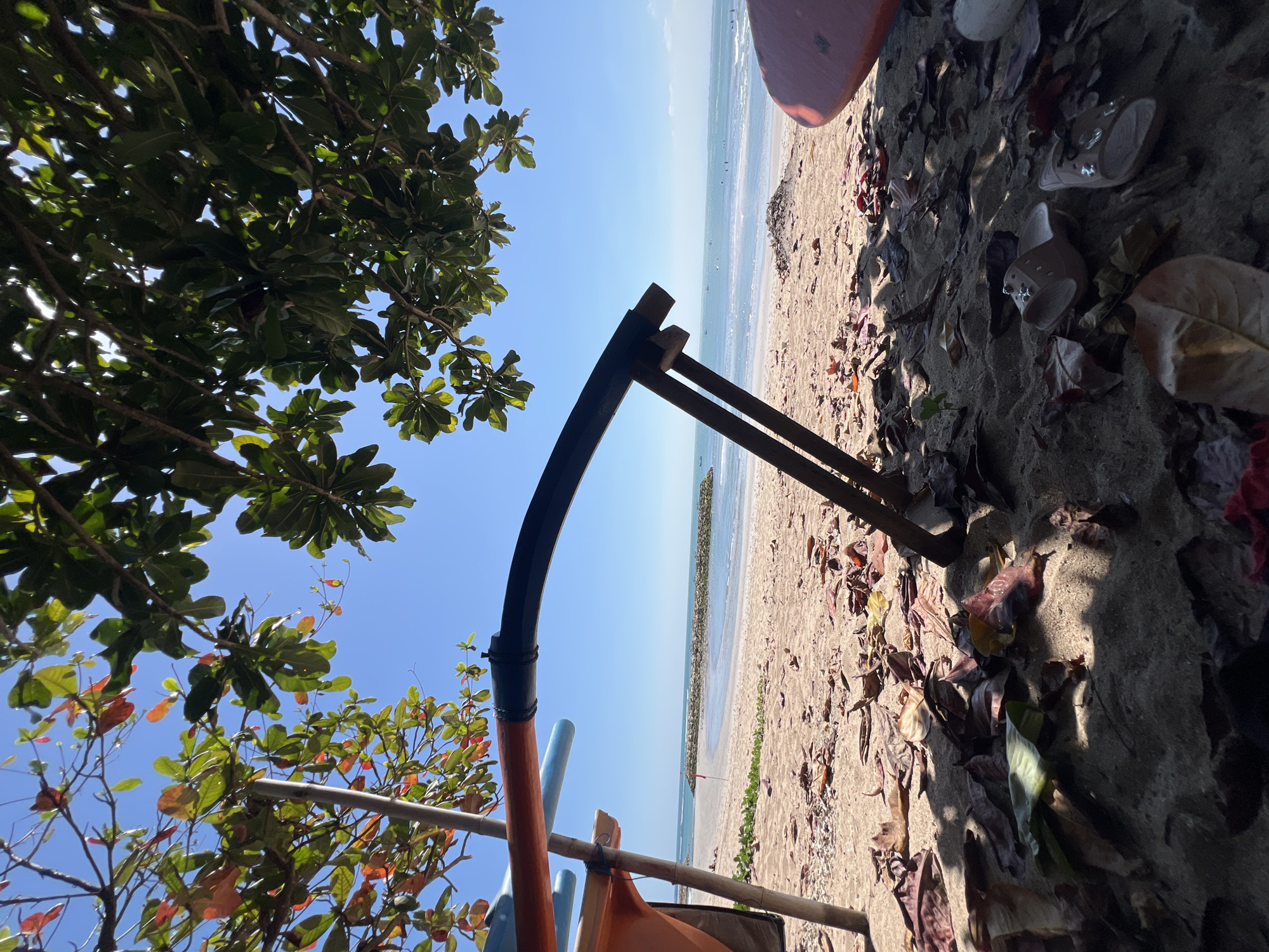 A low-angle, ground-level perspective captures a sandy beach in Kuta, Indonesia, under the shade of a leafy tree. In the foreground, fallen leaves litter the sand, with a pair of light brown flip-flops visible. A portion of an orange and blue outrigger canoe or boat is partially visible on the left, supported by bamboo poles. A dark, curved object, possibly part of a boat's structure, arches across the frame, with wooden supports holding it in place. The background reveals the calm ocean with gentle waves, a distant breakwater made of rocks, and a small red flag. Several small boats and people are scattered in the water. The sky is a clear, bright blue.