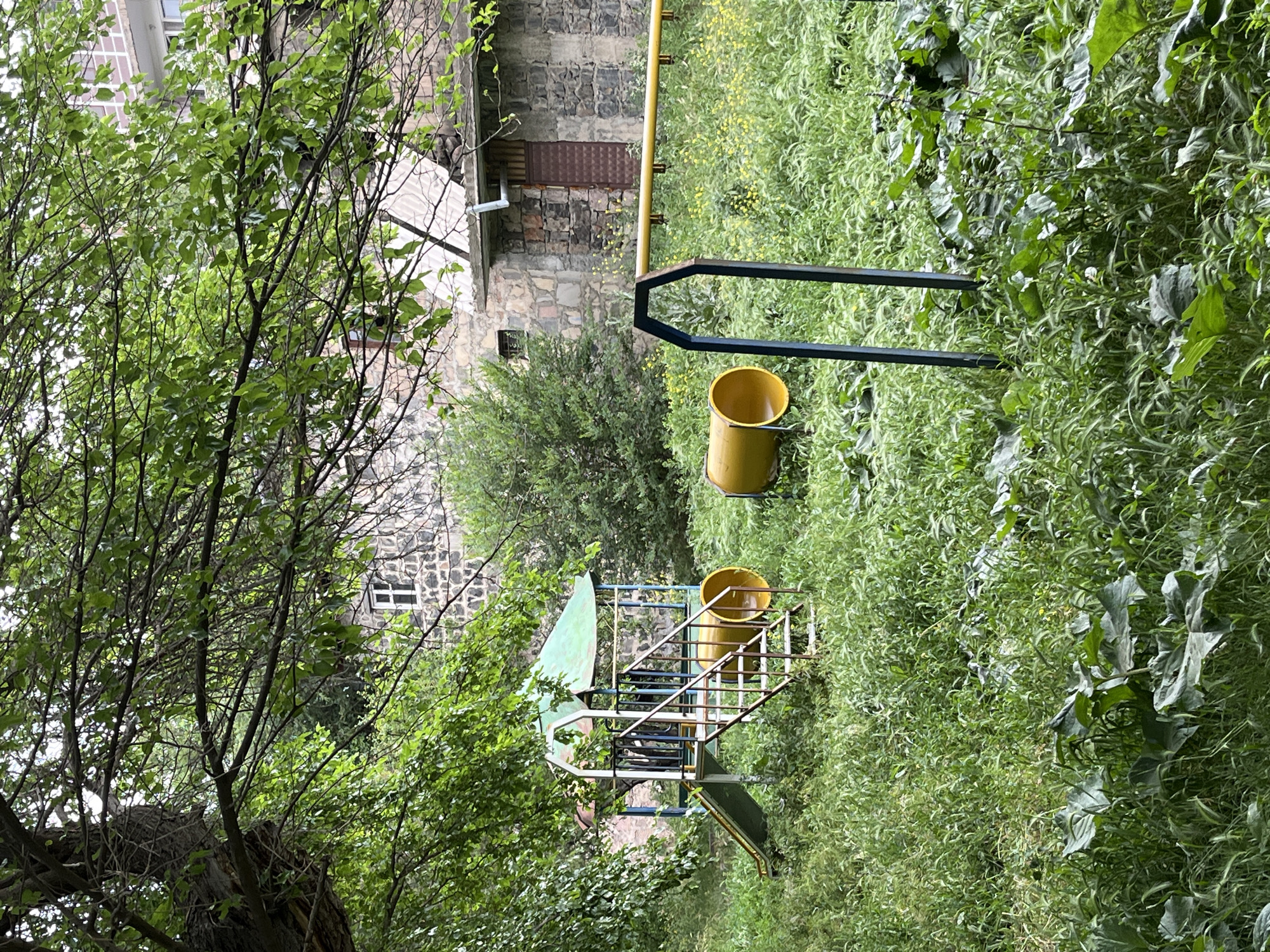 In an overgrown outdoor area, a weathered playground structure stands amidst lush, tall green grass and scattered yellow wildflowers. The playground features a green-roofed fort with a blue and yellow frame, accessible by a metal staircase. A yellow slide extends from the fort, and two large yellow cylindrical tunnels are positioned on the ground in front of it. A blue metal A-frame supports a long yellow horizontal bar. Behind the playground, a stone building with a light-colored roof and a dark brown door is visible, partially obscured by trees. The scene suggests an abandoned or neglected play space in a rural or semi-rural setting.