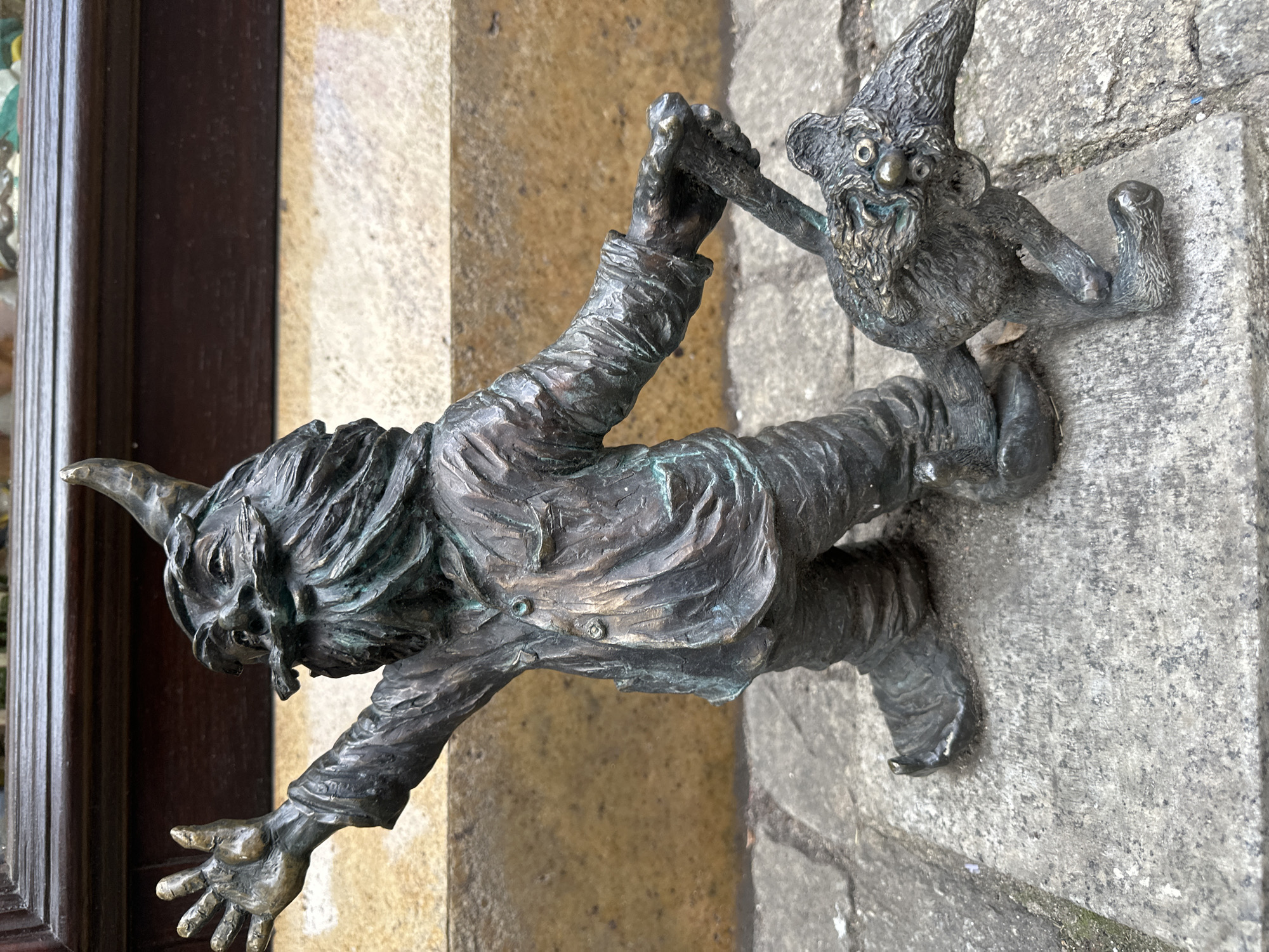 A bronze sculpture of two Wrocław dwarfs is mounted on a building facade. One dwarf, clad in a buttoned jacket, is positioned horizontally, attempting to climb onto the building. His left hand is outstretched, while his right hand grasps the hand of another dwarf perched on the wall. The second dwarf has a pointed hat, beard and distorted facial features. The building facade consists of stone and wood.