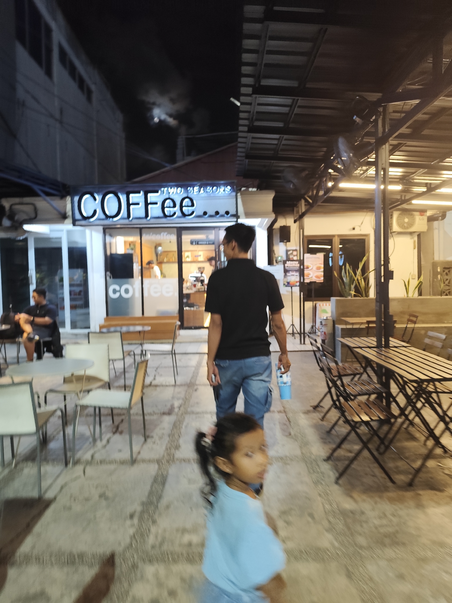 This media file captures an outdoor scene at night, likely a cafe or restaurant. The primary subjects are a man walking away from the camera and a young girl in the foreground, also looking away. The man is wearing a black t-shirt and blue cargo pants, holding a light blue cup. The girl is wearing a light blue t-shirt.
The setting appears to be a paved walkway in front of a building with a prominent neon sign that reads "COFFEE TWO SEASONS". The cafe has large glass windows, revealing a counter and a person inside. There are tables and chairs scattered across the walkway, some set up for patrons and others folded or stacked. Overhead, a dark roof structure with industrial-style lighting is visible. The overall atmosphere is dimly lit, typical of an evening or night setting. The sky above is dark with some illuminated clouds, possibly indicating moonlight or atmospheric conditions.
The image seems to be taken candidly, with the subjects not posing for the camera. The motion blur on the girl suggests movement. There are no clear indicators of specific weather conditions, but the lighting and darkness suggest it is not raining. The scene conveys a casual, relaxed evening outing, possibly at a popular local spot given the cafe's signage. The text "COFFEE" is prominently displayed on the building facade.
