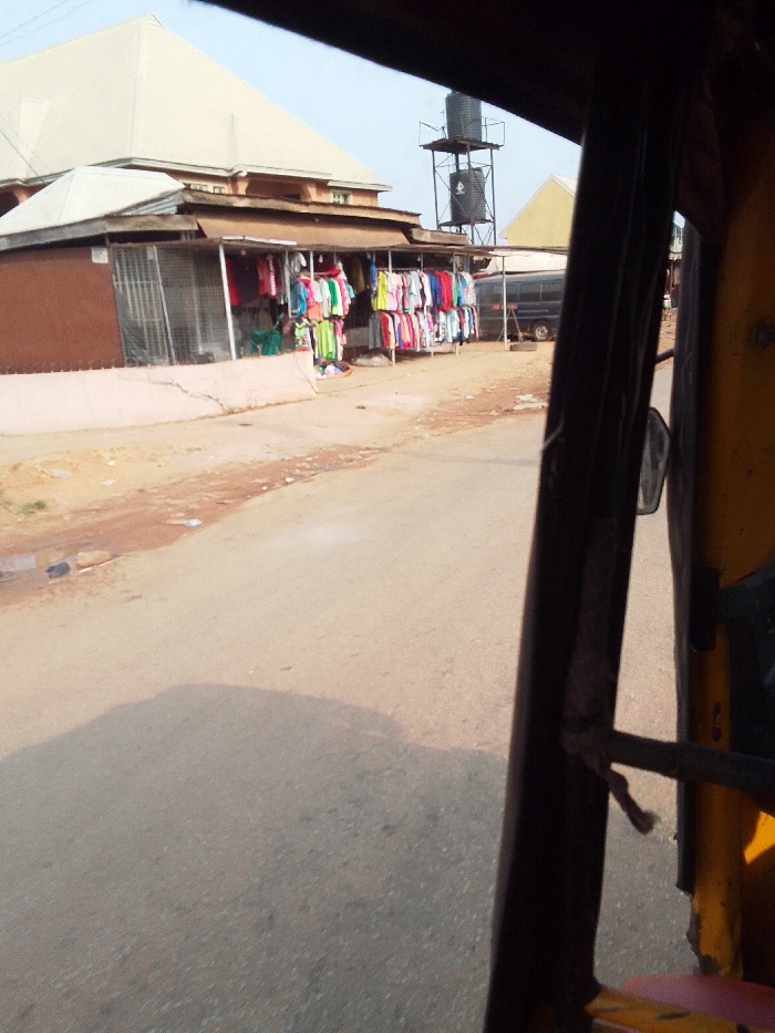 This image presents a street view in Nkwelle Ezunaka, Nigeria, taken from inside a vehicle, likely a tricycle, indicated by the dark frame and yellow interior component on the right. The scene centers on a paved road with minor debris along its left edge. On the left, an open-front shop operates, displaying numerous colorful garments, including t-shirts, blouses, and dresses, hanging on racks. A figure wearing a light green garment is partially visible within the shop. Behind the shop, a multi-story building with a light-colored, sloping roof is prominent. Further down the street on the left, a tall metal structure supports two black water tanks against a clear sky. A dark-colored minivan or bus is parked on the right side of the street, facing away from the viewer. A vehicle's shadow is cast diagonally across the paved road surface in the foreground.