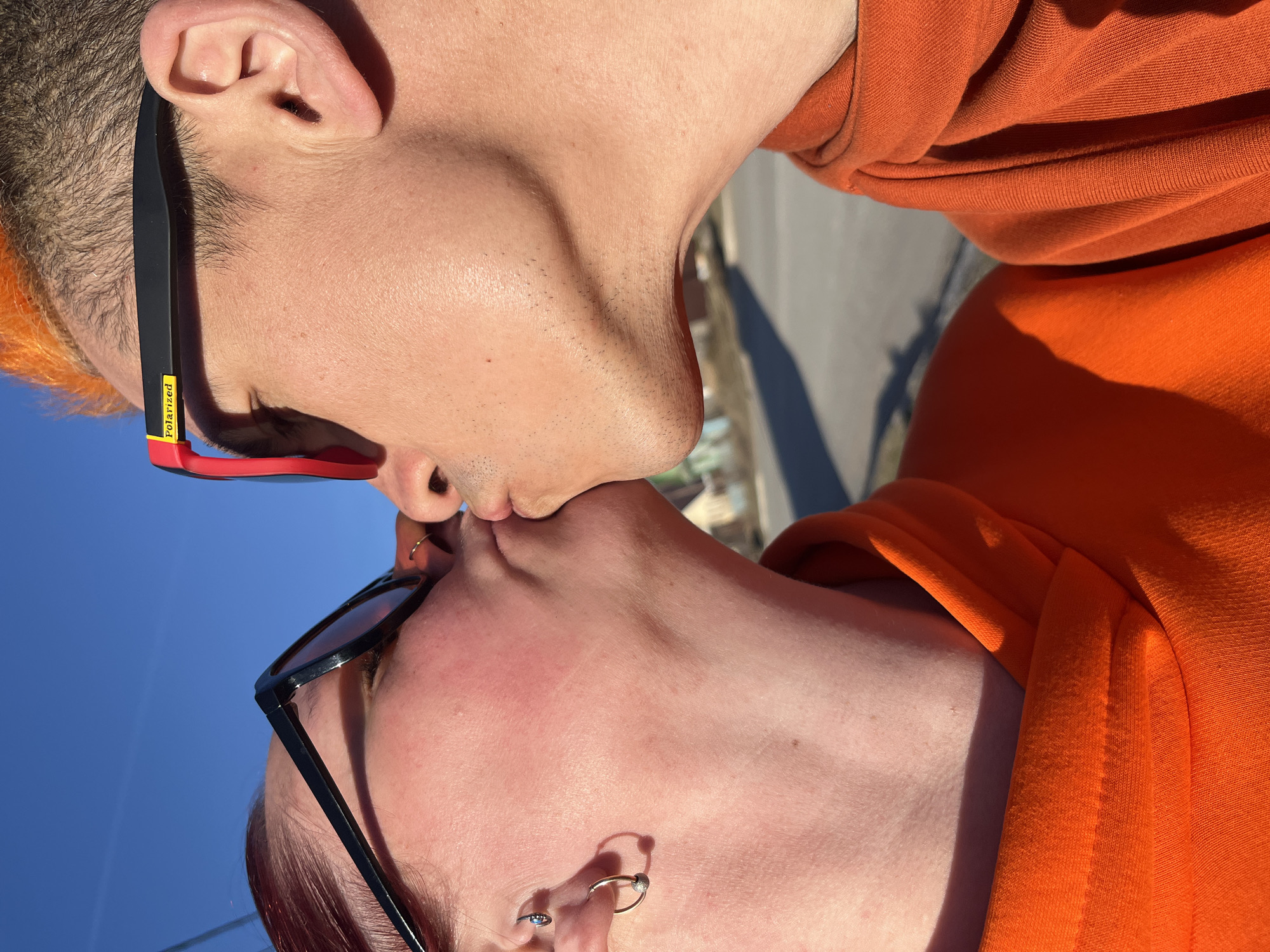 This image captures a close-up, side-profile view of two individuals kissing outdoors in Chaikovsky, Russia, against a bright blue sky. Both subjects are wearing orange hoodies and sunglasses, and their heads are tilted towards each other to meet in a kiss.
The person on the top has short, spiky orange hair and is wearing black sunglasses with red accents on the arms. The word "Polarized" is visible in yellow text on the temple of their sunglasses. Their skin shows some light stubble on the chin and jawline.
The person on the bottom has red hair and is wearing black sunglasses. They have multiple ear piercings, including a prominent hoop and a stud, and a small nose ring is visible.
The scene is brightly lit by the sun, casting distinct shadows on their faces and clothing, highlighting the texture of the orange hoodies. The clear, vibrant blue sky dominates the background, with some blurry, indistinct structures or trees visible in the far distance, suggesting an outdoor setting during the daytime with good weather. The image conveys an intimate and affectionate moment between the two subjects.