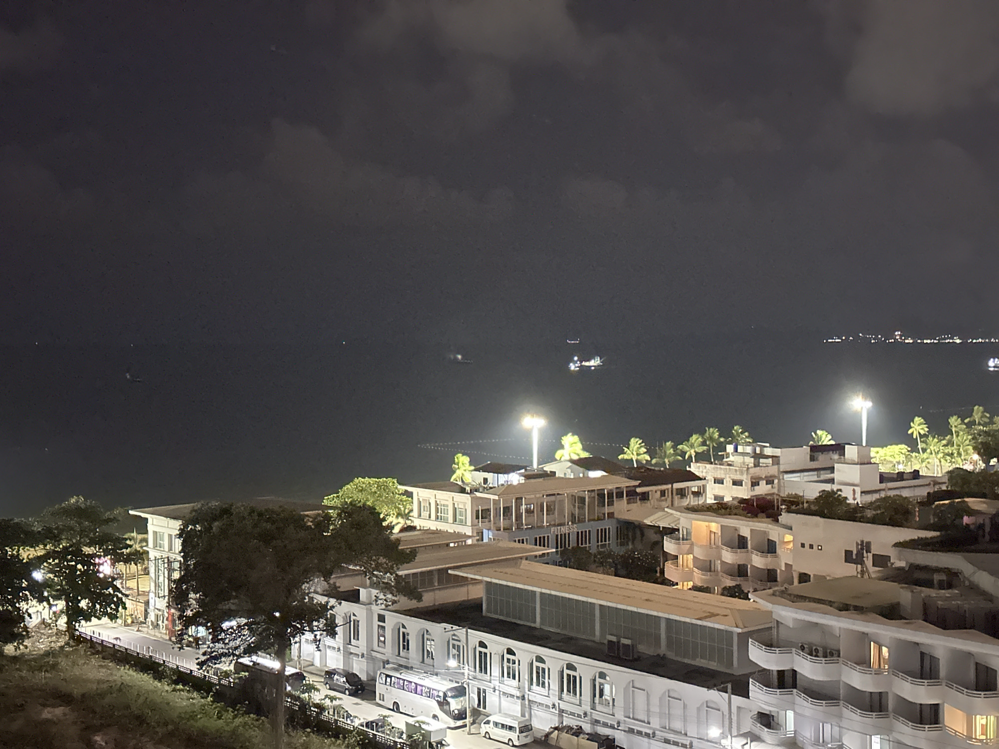 This image is a nighttime view of Pattaya City, Thailand. The scene includes a body of water, likely the sea, with a few lights visible in the distance on the horizon. Closer to the foreground, there are several buildings lit up, some with palm trees visible in the vicinity. In front of the buildings is a street with vehicles parked along it. The sky is overcast with dark clouds.