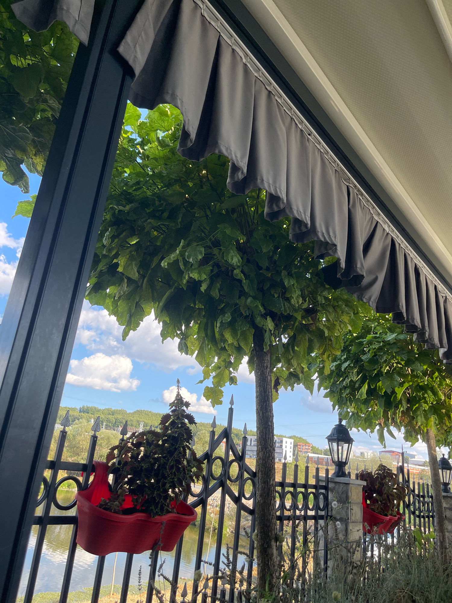 The image depicts an outdoor seating area with a view of a body of water and a treelined landscape. In the foreground, a grey awning is visible, partially obscuring the lush green leaves of trees. A black metal fence runs horizontally across the scene, adorned with decorative elements. Several red planters containing dark-leaved plants are attached to the fence. A black lamppost stands on a stone pillar, also decorated with a planter. In the background, buildings and trees are visible on a hill overlooking the water. The sky is blue with scattered white clouds. The scene appears to be in Mirushë, Kosovo, suggested by the presence of a lake, hills, and modern buildings characteristic of the area.