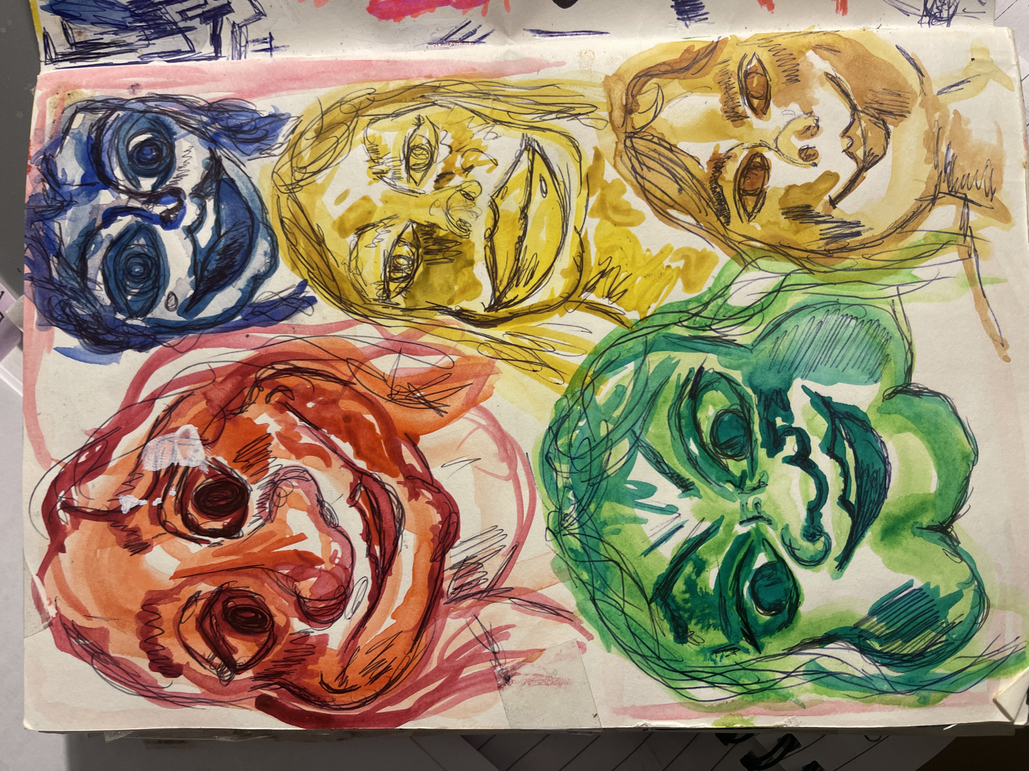 The image shows a page from a sketchbook. On the page, there are five abstract, stylized face drawings, each rendered in a different color: blue, yellow, light brown/tan, red/orange, and green. The faces are positioned upside down. Each face is outlined with a dark line and filled with color washes. The colors are vibrant and bleed slightly outside the drawn lines. The style is loose and expressive, with visible sketching and layering. The background of the page is white. The upper left corner of the page has some blue scribbles. There are no clear indicators or associations to suggest that this image depicts Maidstone, United Kingdom.