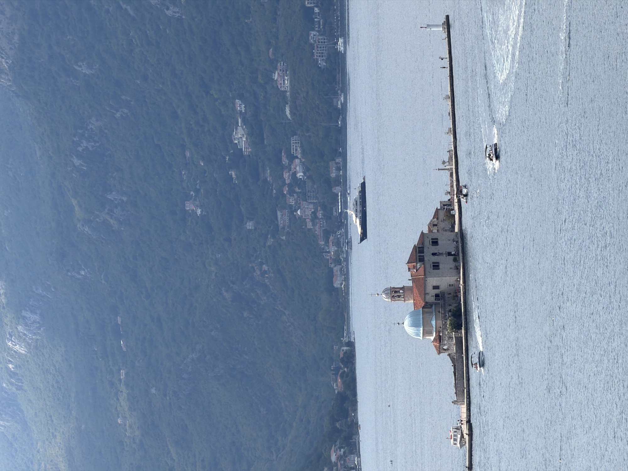The image captures the iconic island church of Our Lady of the Rocks, a man-made islet located in the Bay of Kotor, near the historic town of Perast, Montenegro. The church, with its distinctive blue dome and red-tiled roof, sits on a small, rocky island connected by a stone walkway to a larger section of the islet. Several boats are visible in the surrounding tranquil, blue water. In the distance, the steep, verdant slopes of the surrounding mountains rise, dotted with scattered houses, indicating the proximity of the town of Perast. A large yacht is also seen sailing further out in the bay. The overall scene is serene and picturesque, highlighting the unique architectural and natural beauty of the location.