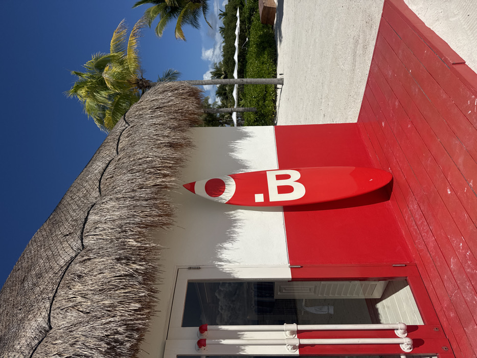 This image captures an outdoor scene at Marina Maroma, Mexico, under a clear blue sky. A building with a natural thatched roof forms the primary structure, its facade horizontally divided into an upper white section and a lower bright red section. A white-framed glass double door is situated on the left, reflecting ambient light. To the immediate left of the door, three white, vertical pipe-like structures are mounted. A bright red surfboard, approximately two meters in height, leans against the red wall section, displaying a prominent white circular design above a capitalized white letter "B." A red wooden deck extends from the base of the building, transitioning onto a white sandy ground. In the background, multiple palm trees with green fronds are visible, alongside lush green foliage and several distant white umbrellas or shade structures. The scene is brightly illuminated by direct sunlight, evidenced by distinct shadows cast by the roofline and the surfboard. No individuals or animals are present.