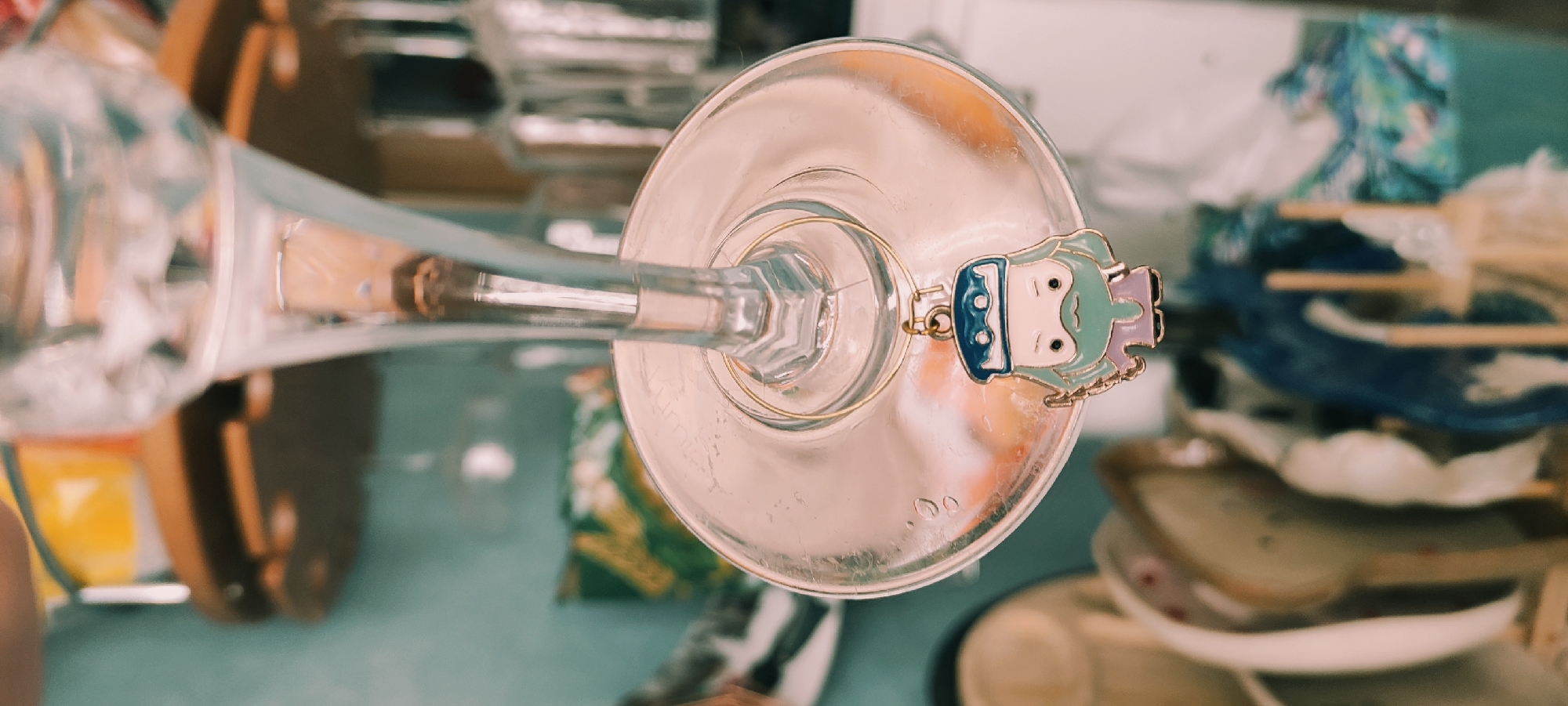 An overhead shot depicts a clear glass object resembling a champagne flute, viewed from above, and lying horizontally. A metallic ring with a blue and mint-green enamel charm of a stylized child-like figure hangs from the rim of the flute. The flute sits on a surface with a teal or pale blue cast. The background features a cluttered assortment of items, including what appear to be wooden trays, a blue bowl or plate, fabric, and boxes, all in a slightly out-of-focus state.