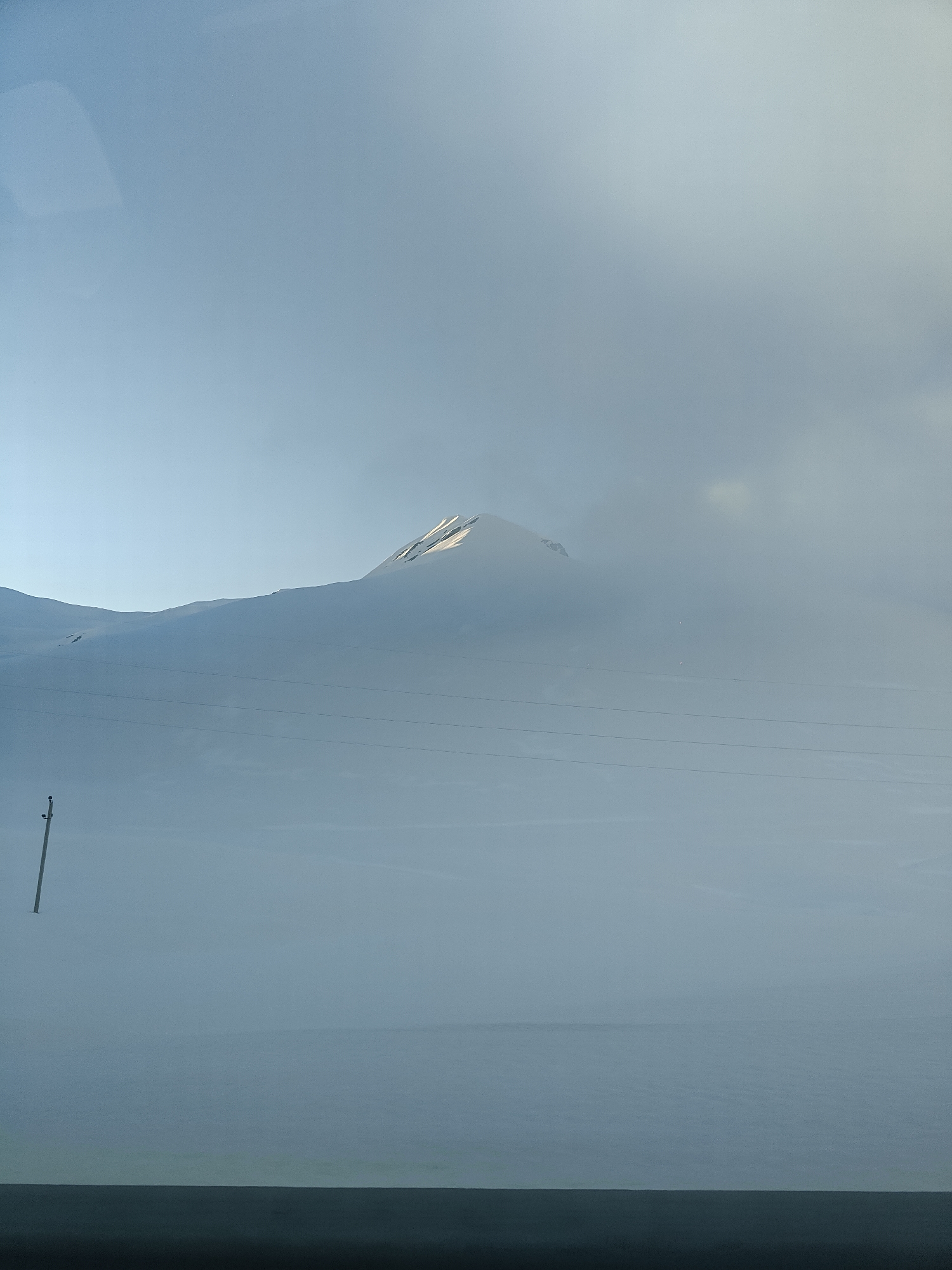The image captures a vast, snow-covered mountainous landscape in Ganisi, Georgia. The scene is dominated by a blanket of white snow, covering rolling hills and mountain slopes that extend into the distance. A faint, distant mountain peak is visible towards the center-right, its uppermost section catching soft sunlight, suggesting either early morning or late afternoon.
The sky is a pale, hazy blue, transitioning into a lighter, cloud-filled expanse that blends seamlessly with the snow-covered terrain, particularly in the mid-ground, where atmospheric fog or low clouds obscure parts of the view. In the left foreground, a tall, dark utility pole stands upright, emerging from the deep snow. Multiple dark power lines stretch horizontally across the middle of the frame, disappearing into the distant haze.
The overall impression is one of serene isolation and immense scale, characterized by the cold, white expanse. No people or visible text are present. The dark band at the very bottom of the image suggests the photograph was taken from inside a vehicle.