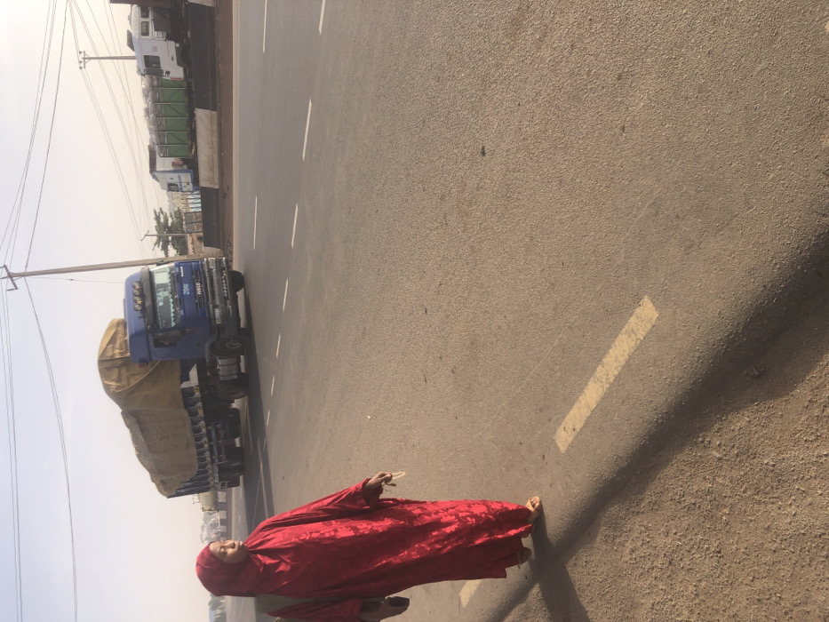 The image depicts an outdoor scene on a paved road in Hanwa, Nigeria, under a hazy, light-colored sky. In the foreground on the left, an individual, appearing female, walks on an unpaved, reddish-brown dirt shoulder. She is dressed in a long, red, patterned garment and a matching red hijab covering her head and neck. Light-colored sandals are visible on her feet. A dark bag is carried on her left shoulder, and she holds a small, light-colored object in her right hand. Her gaze is directed towards the right side of the frame.
Adjacent to the unpaved shoulder is a raised curb with a yellow dashed line, bordering a multi-lane asphalt road marked with white dashed lane lines. A blue Iveco commercial truck, consisting of a tractor unit pulling a trailer covered with a light brown tarpaulin over a significant load, is observed moving from right to left on the paved road in the middle ground. In the far background on the right, several other commercial trucks, including white and green models, are stationary or parked on an adjacent paved area. A utility pole with power lines is visible on the right side of the frame, extending upwards above the road. Distant, indistinct structures are faintly visible through the atmospheric haze.