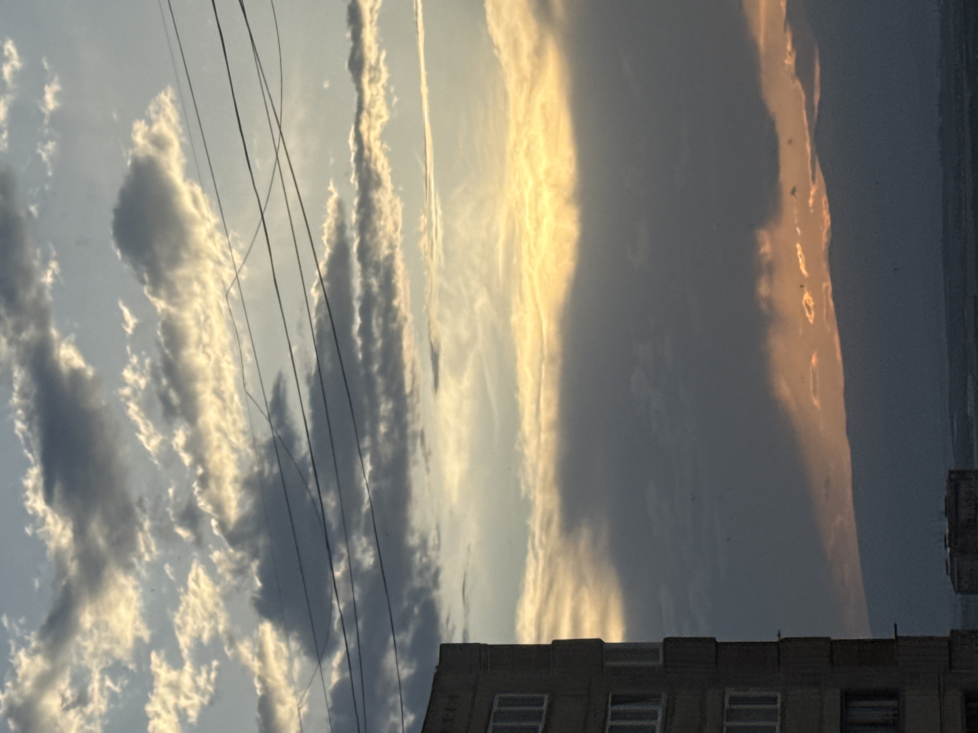 This is a vertical, eye-level, medium shot of a city skyline in Abovyan, Armenia, under a cloudy sky at either sunrise or sunset. The left side of the frame is dominated by a portion of a tall, concrete, apartment building. The building has multiple visible windows. Several dark, overhead wires crisscross the sky in the upper half of the image. The sky is filled with clouds that range from light grey to dark grey, with patches of bright yellow and orange light breaking through, indicating the presence of the sun. In the background, a range of dark, silhouetted mountains is visible, extending across the bottom portion of the image. A lower, dark-colored building with a flat roof is visible in the distance. The overall impression is one of an urban environment at dusk or dawn.