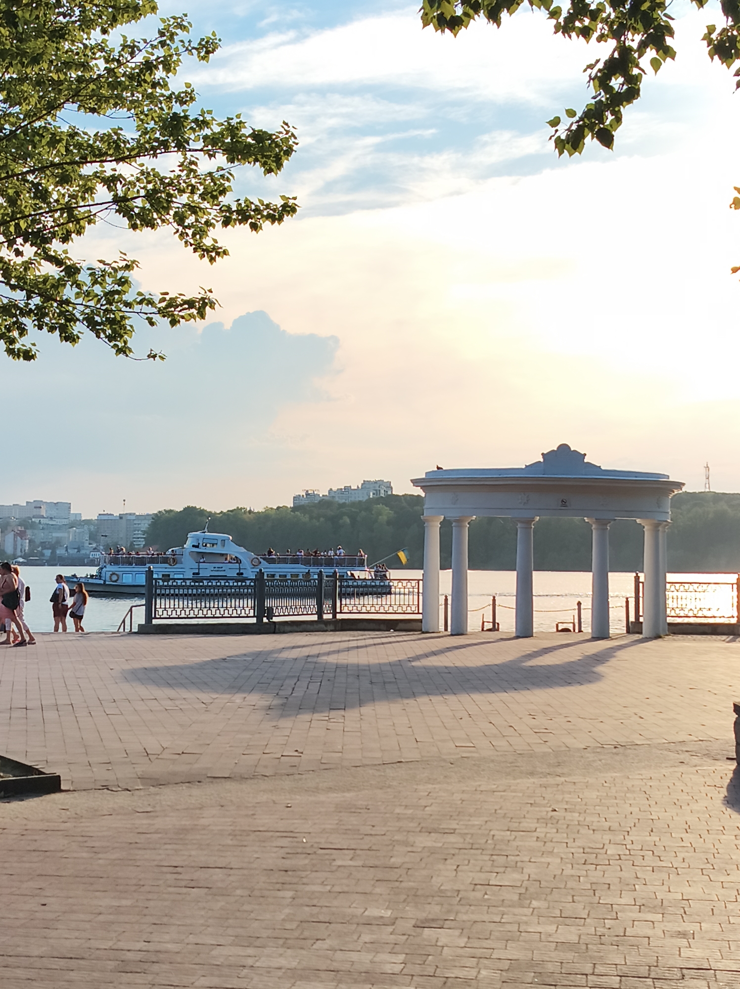 The image captures a scenic view of the Ternopil lake embankment in Ternopil, Ukraine, during what appears to be a sunset. In the foreground, a paved walkway with a distinct brick pattern stretches across the frame, with long shadows cast by an architectural structure. This structure, a white rotunda with multiple columns and an ornate roof, stands prominently on the right side of the image, overlooking the water. On the left, a white passenger boat with a crowded deck, filled with people, is docked near the embankment. Several individuals are walking along the walkway in the mid-ground, some appearing to be tourists. The background reveals a cityscape with buildings and trees, nestled against a bright, hazy sky with scattered clouds, indicating warm weather conditions. The overall atmosphere is serene and picturesque, typical of a recreational urban waterfront.