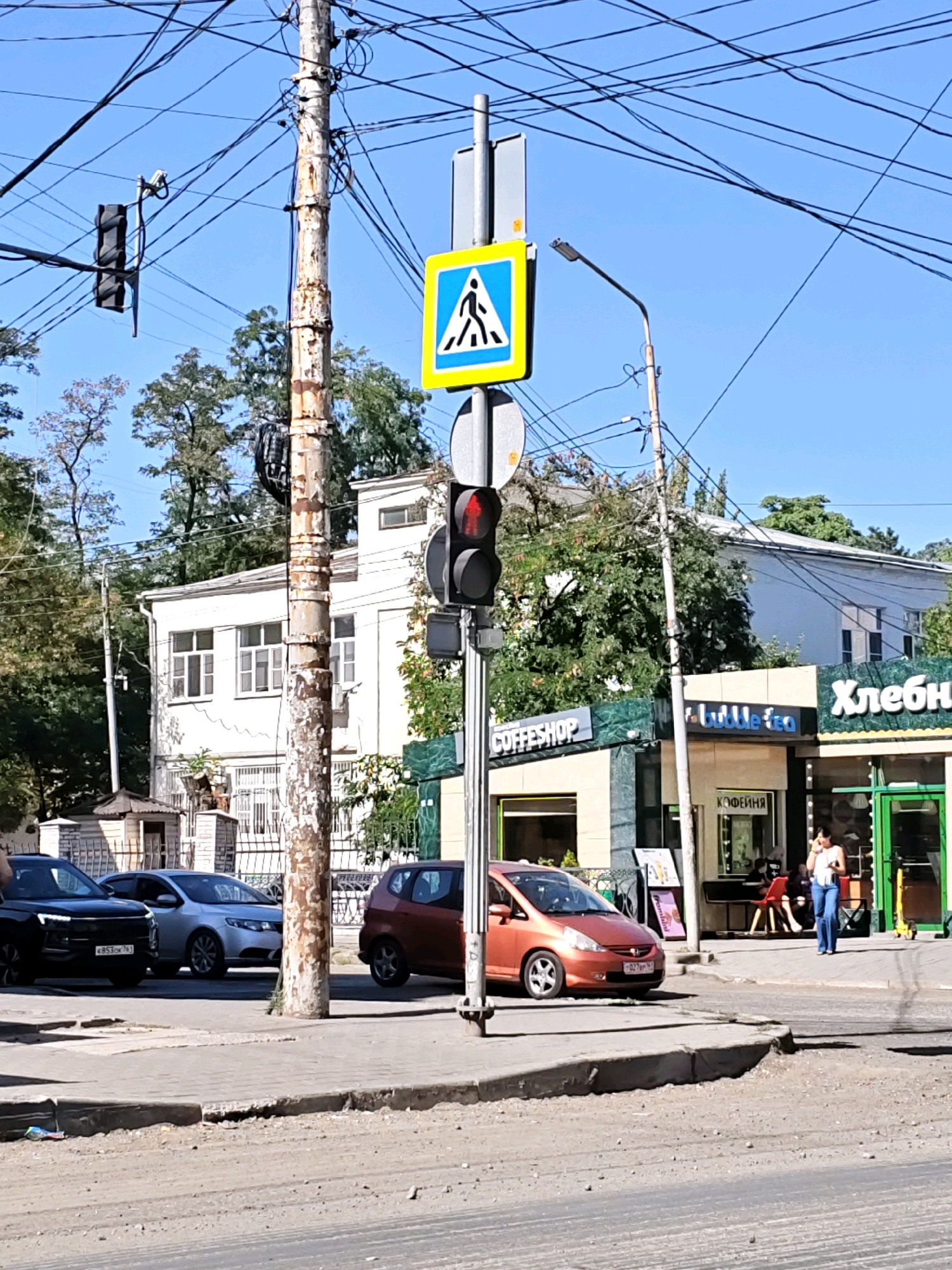 The image shows a street scene in Rostov-on-Don, Russia. In the foreground, a dirt road is visible with a concrete curb. To the left, several cars are parked on the side of the road, including a black SUV and a silver sedan. A pedestrian crossing sign with a blue background and a white figure is displayed prominently. A traffic light with red illumination is visible. Behind the cars, a white building with multiple windows stands tall. In front of the building, there are shops with green awnings. One shop displays the word "COFFESHOP" in English, and another displays "Хлебная" in Russian, indicating a bakery. A woman is walking on the sidewalk in front of one of the shops, talking on her phone. The environment is characterized by a clear blue sky, trees, and a utility pole with numerous wires. The overall scene depicts a typical urban environment in a Russian city.