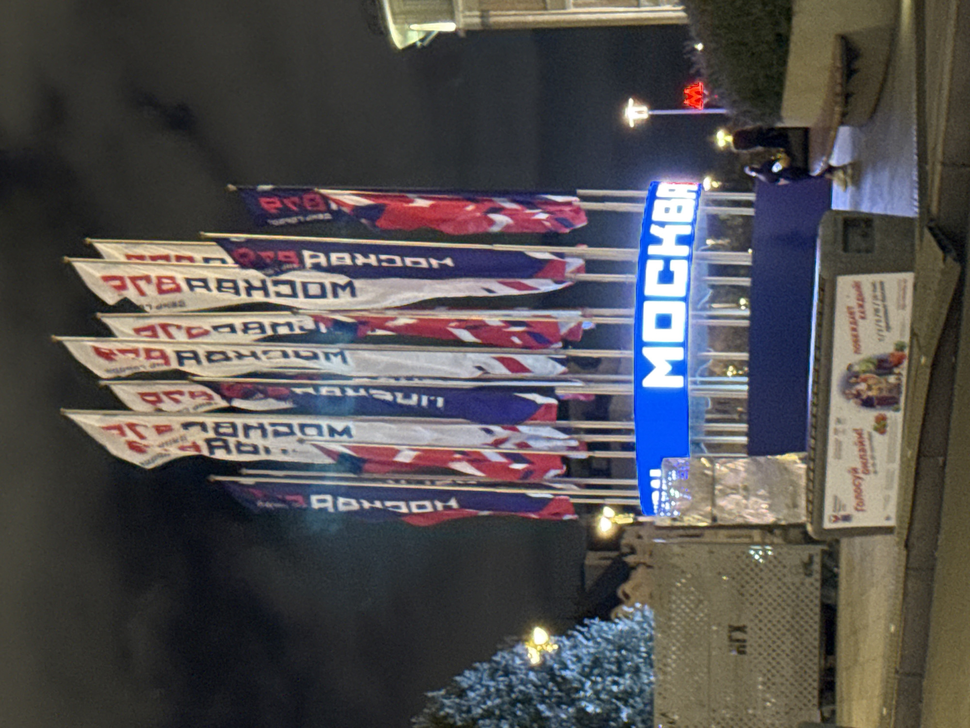 The image shows a street scene at night in Moscow, Russia, featuring a prominent blue illuminated sign with the word "МОСКВА" (MOSCOW) in white lettering. Several large flags are displayed behind the sign, most of which are white with red and blue lettering. Some of the flags feature repeating patterns of red and blue stripes. In the background, a densely decorated tree, possibly an artificial Christmas tree given the season, is visible on the left. Streetlights with a warm glow illuminate the scene. Further to the right, a red illuminated sign is visible. Two people are present in the scene; one is seated on a bench to the right, and another person is standing behind them. In the foreground, there is a low-lying structure with posters and informational displays, one of which has text and imagery related to voting. A metal grate structure is also visible on the left. The sky is dark with some cloud cover.