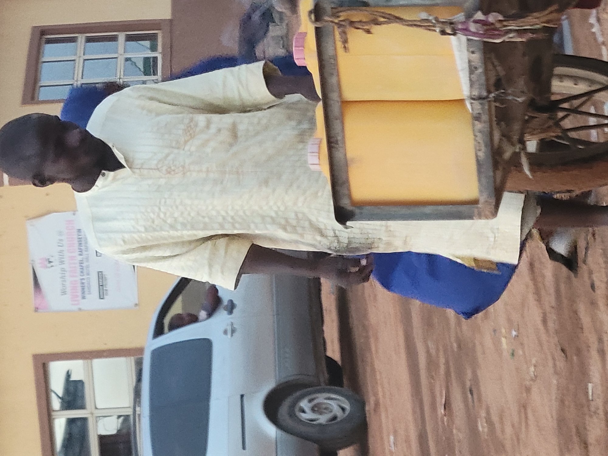 A man in Suleja, Nigeria, is riding a bicycle carrying containers of yellow liquid. The man, who appears to be of African descent, is wearing a light-colored patterned shirt and blue trousers. The containers are loaded in a metal rack attached to the bicycle. In the background, there is a white van parked in front of a building with windows and a poster that reads "Worship With Us, LIVING FAITH CHURCH, WINNER'S CHAPEL, RAFINSEYIN." The ground is reddish-brown, suggesting a dirt or paved surface.