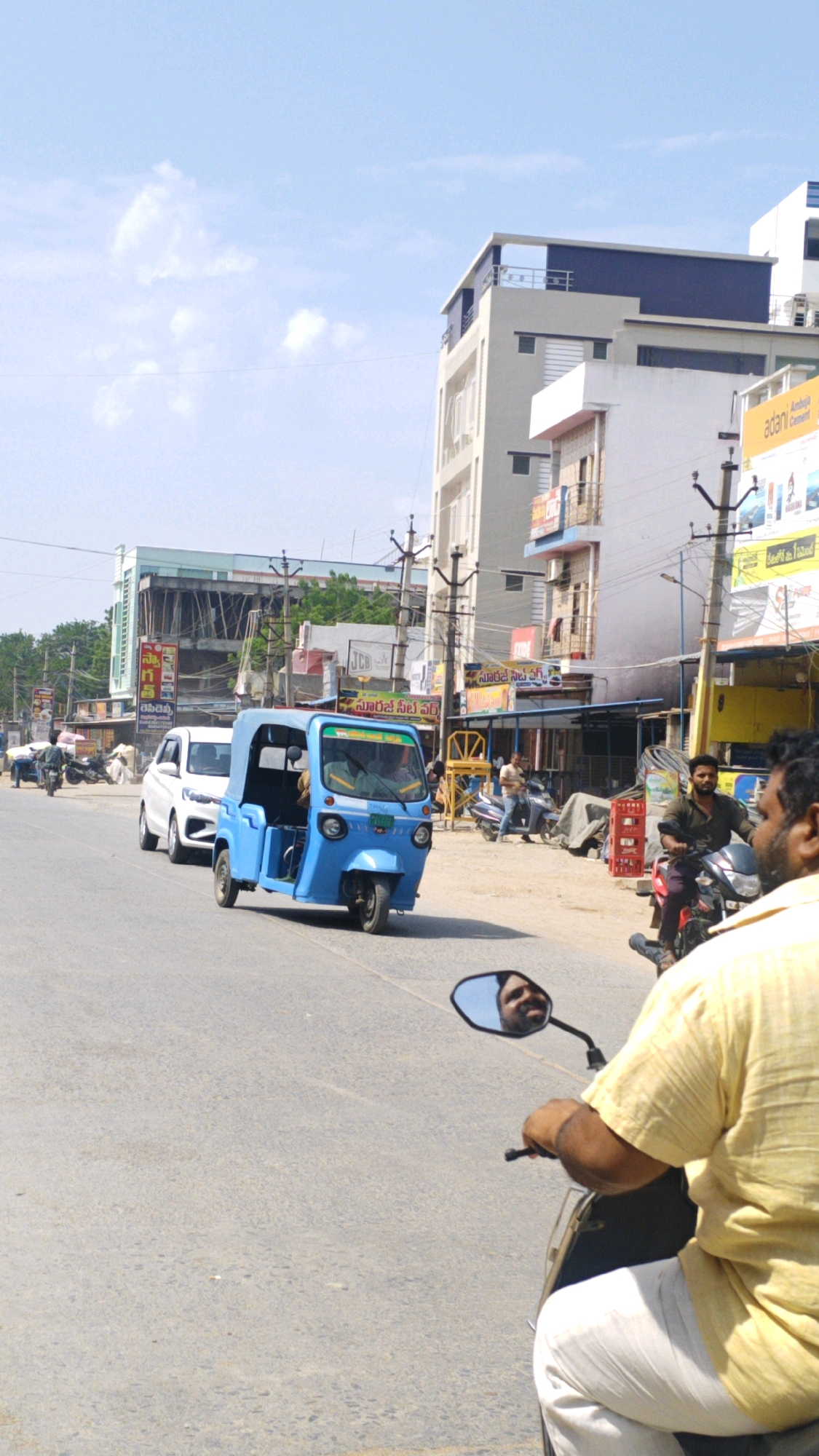The image depicts a street scene in Pamur, India, on a sunny day. The foreground features a person riding a scooter, with their reflection visible in the scooter's side mirror. The person is wearing a yellow shirt and white pants. The scooter is on a paved road. In the mid-ground, a blue auto-rickshaw is driving on the road, followed by a white car. The auto-rickshaw has a canvas cover. A person is visible driving the auto-rickshaw. Further back, various buildings line the street, including a multi-story white building with blue accents and a building under construction with scaffolding. Shops with signage are visible along the roadside. Several other individuals and motorcycles are present on the street. The sky is partly cloudy.