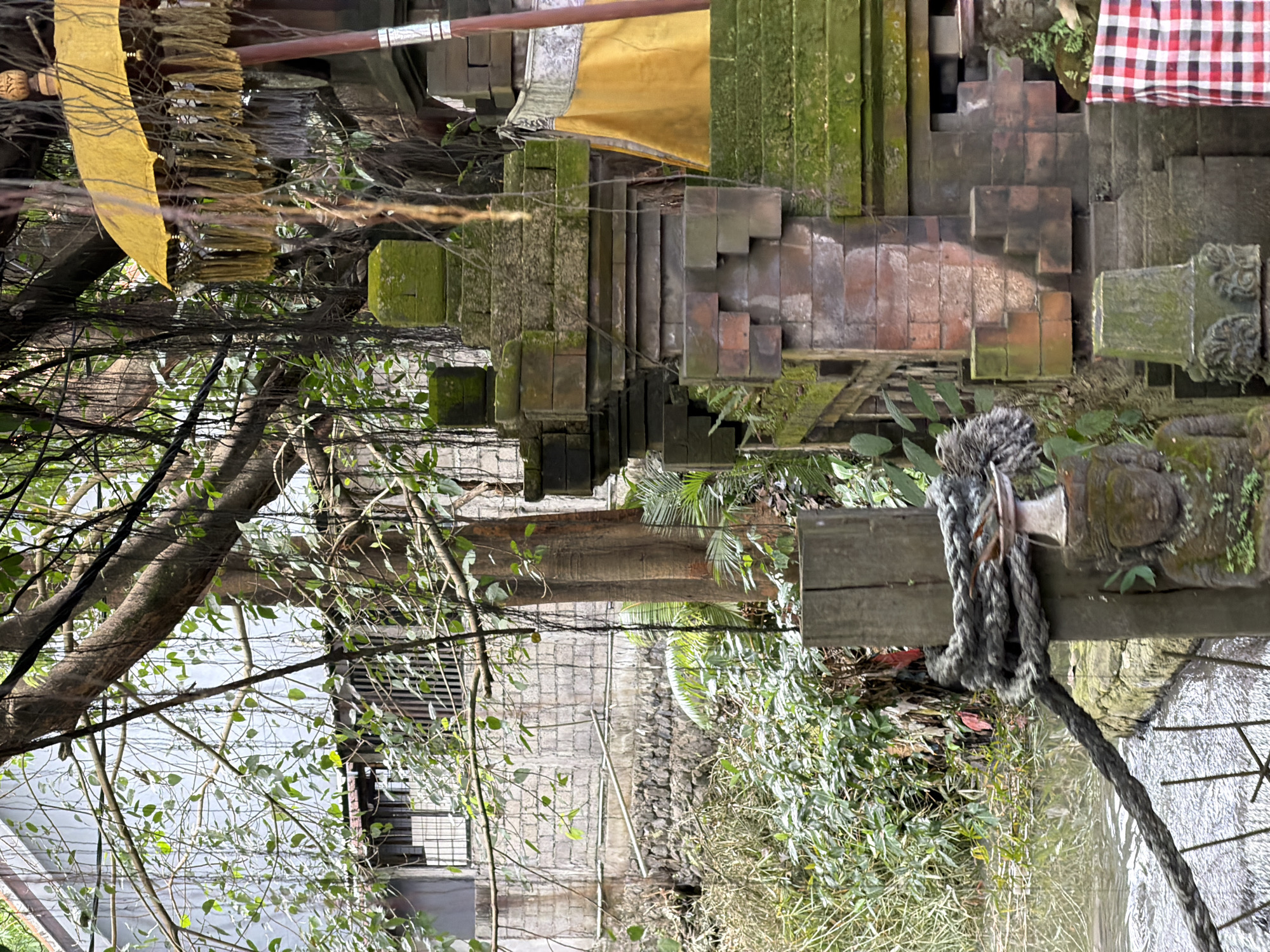 The image depicts an outdoor scene in North Kuta, Indonesia. It features a Balinese temple structure with stone steps covered in moss and greenery. A yellow and white fabric adorns part of the temple. In the foreground, a thick rope is tied to a wooden beam near foliage. Through the branches of trees, the background reveals a building with a grey facade and a wall constructed of dark stones. The perspective is angled, capturing both the temple's architectural details and the surrounding natural environment.