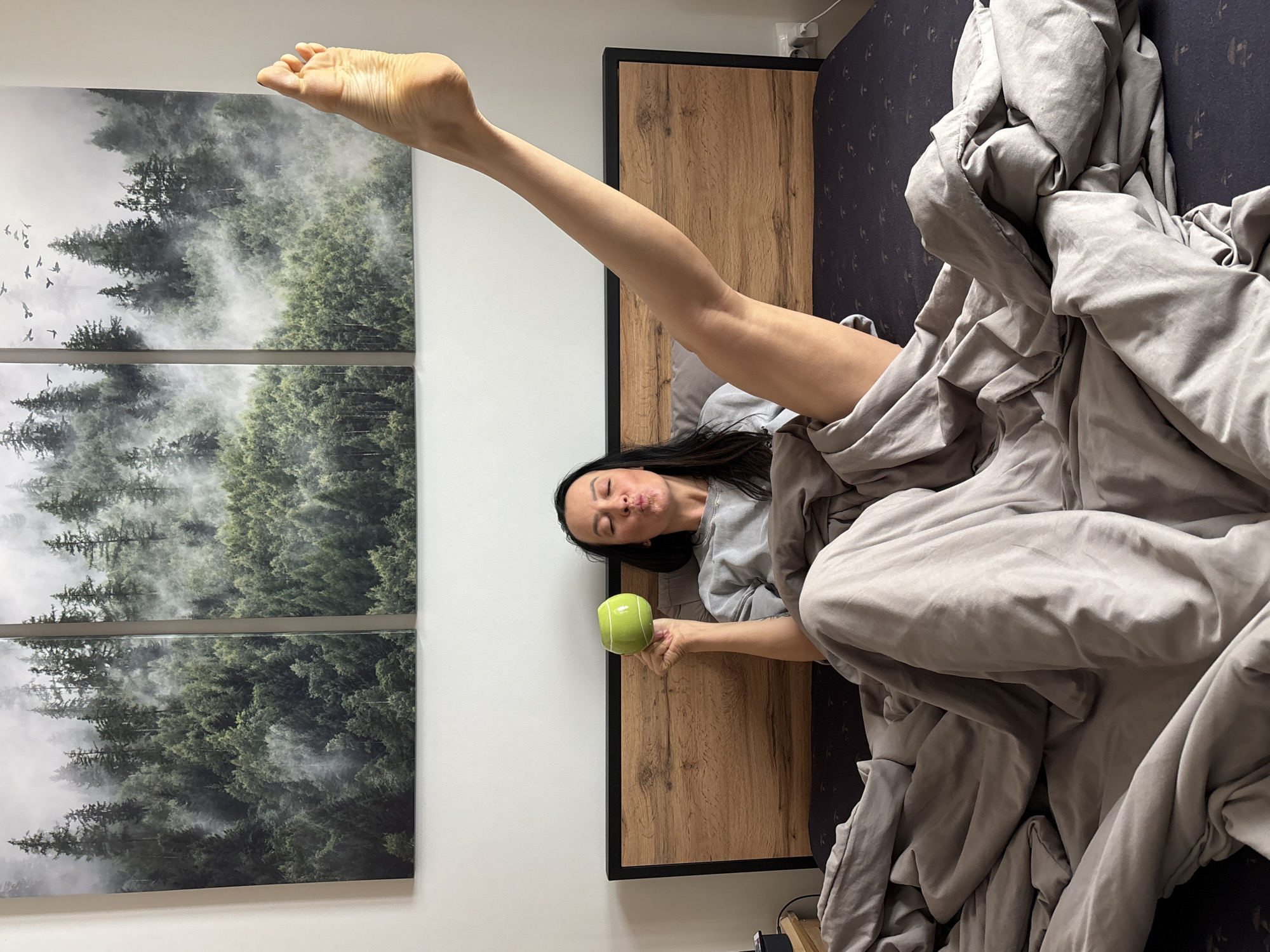 A woman lies in bed under a gray comforter, holding a green mug in her right hand. Her left leg is extended straight up into the air, with her foot near a triptych wall art piece that depicts a forest scene. The headboard is a light wood with a dark border. The location is likely a bedroom in a private residence; the city of Chaikovsky, Russia cannot be definitively determined from the image.