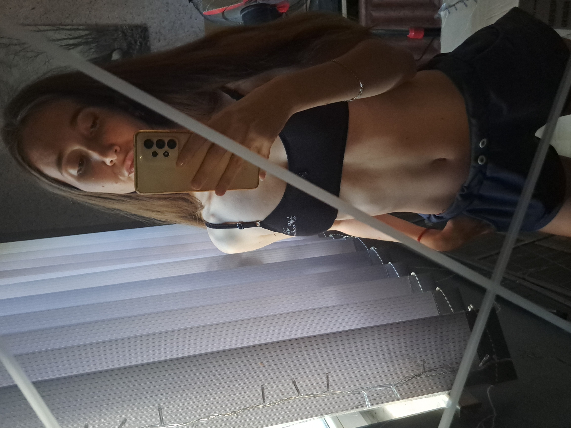 A young woman with long brown hair is taking a mirror selfie in what appears to be a bedroom. She is wearing a black sports bra and dark blue athletic shorts. The room has vertical blinds covering a window on the left side of the frame. There is a string of fairy lights on the left side of the window. The background shows a wall with a framed picture of trees. The woman is holding a gold-colored smartphone to take the picture. The lighting suggests it is daytime.