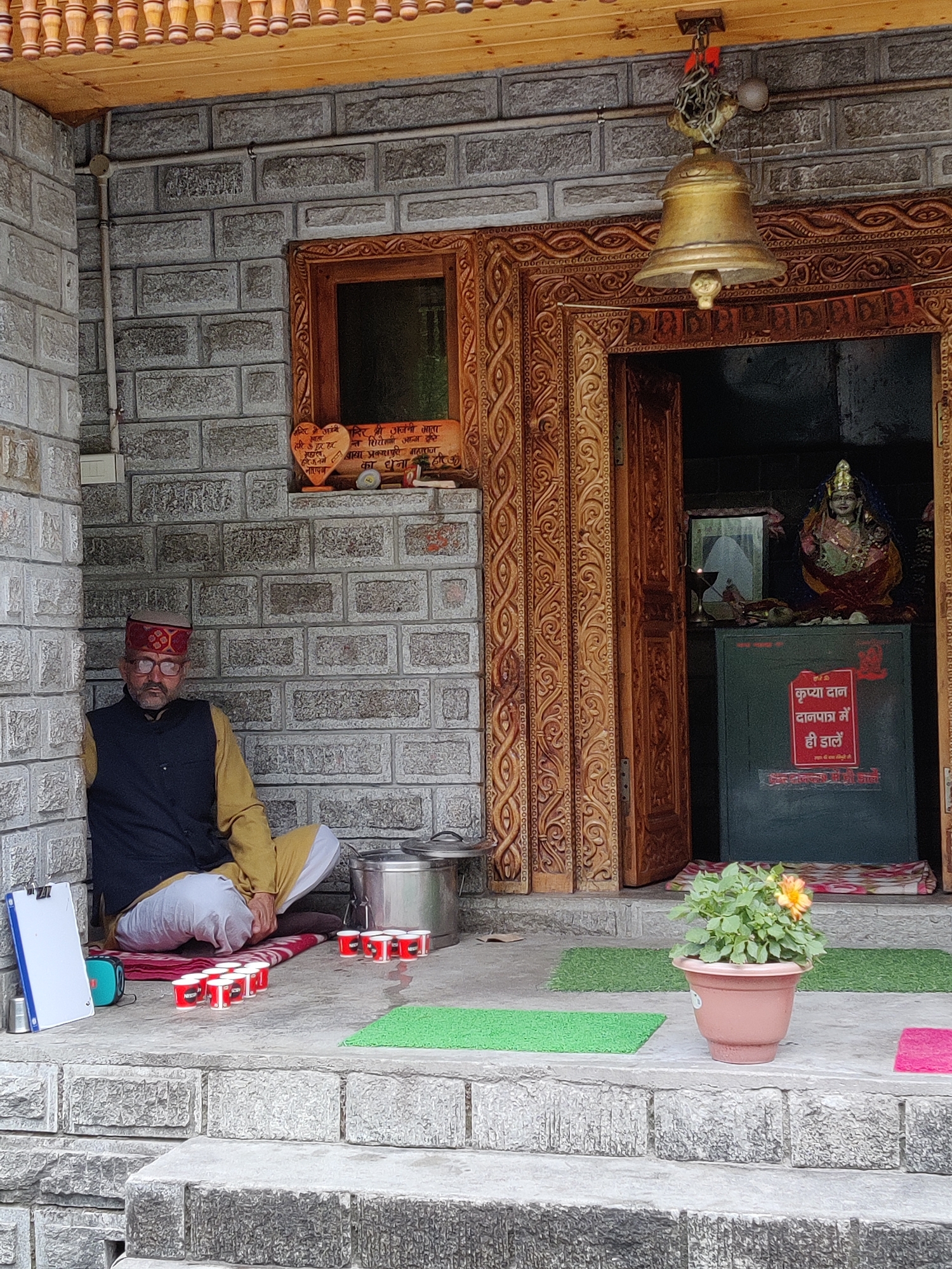 A man in traditional Indian attire, including a red and yellow hat, a dark vest, and yellow and white clothing, sits on a mat in a stone alcove outside a building. He is wearing glasses. In front of him, on the ground, are several small red cups, some with white lids, arranged in rows. A large metal pot and a clipboard are also nearby. To the right of the man, an open doorway leads to a shrine where a statue of a deity is visible, adorned with colorful clothing and jewelry. A bell hangs above the doorway. A potted plant with a single orange flower sits on the steps leading to the entrance. The building's exterior is constructed with grey bricks, and the doorway features intricate wooden carvings. A sign with Hindi text is affixed to a green box inside the shrine. The scene appears to be an outdoor setting in a village or town, possibly a temple or a place of worship.