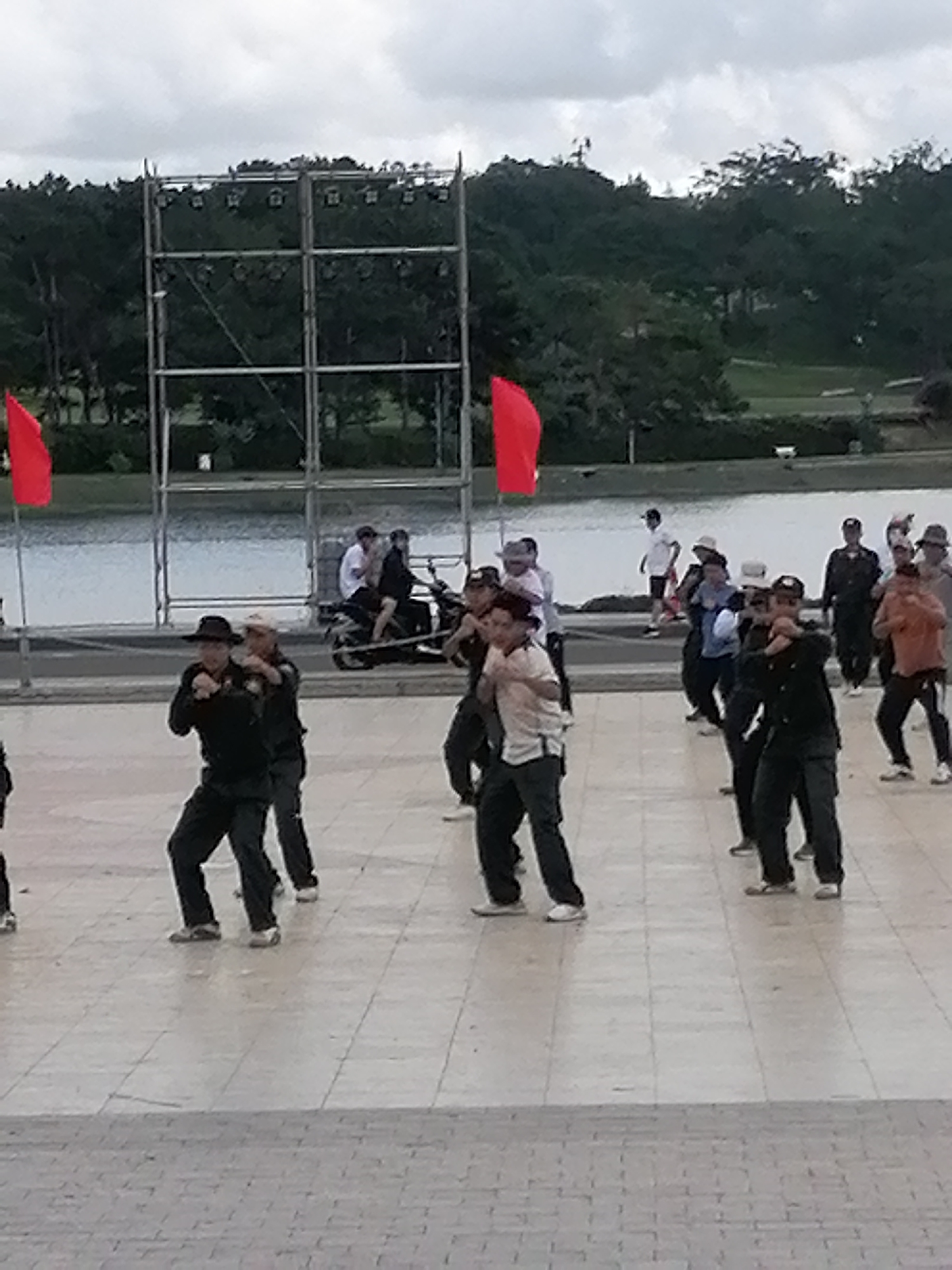 In an outdoor, paved plaza, a group of people are engaged in what appears to be a martial arts or self-defense class. The individuals, dressed in dark clothing and some in hats, are positioned in various stances with their fists raised. Some are in the foreground, actively participating, while others are further back. In the background, a body of water is visible, with a line of trees and a cloudy sky above. A metal structure, possibly a stage or lighting rig, stands on the left side of the image, with a red flag attached. A motorcycle with two people on it is also present in the mid-ground. The overall atmosphere suggests a public demonstration or training session in an urban park setting.