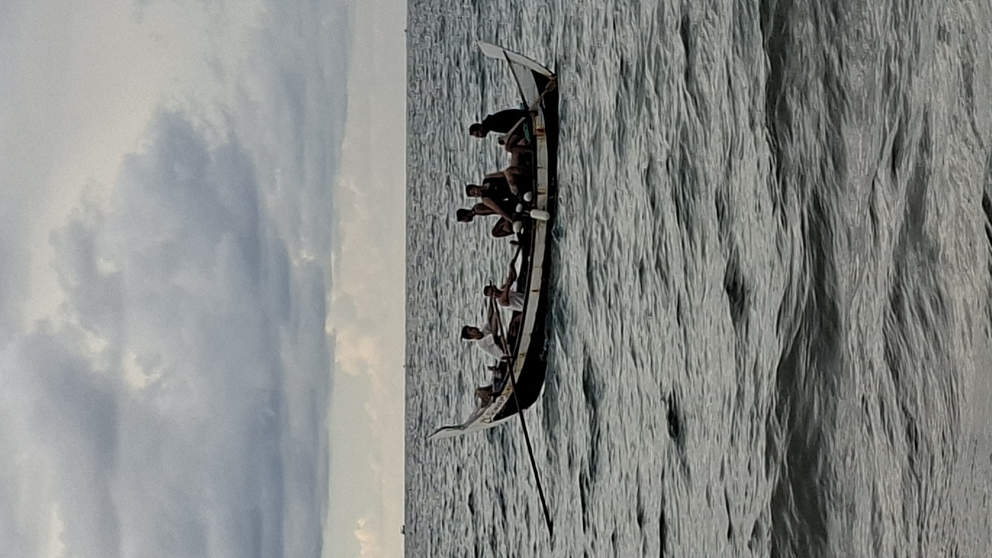 A long, narrow boat with a dark hull and white trim is moving through choppy, dark gray water under an overcast sky. Six men are seated in the boat. Two men in the front are rowing with oars. The other four men are seated further back in the boat. The boat is moving from left to right in the frame. The water is reflecting the dark sky, creating a somber mood. The scene is consistent with the waters off the coast of Lhokseumawe, Indonesia, where such traditional fishing boats and overcast skies are common.
