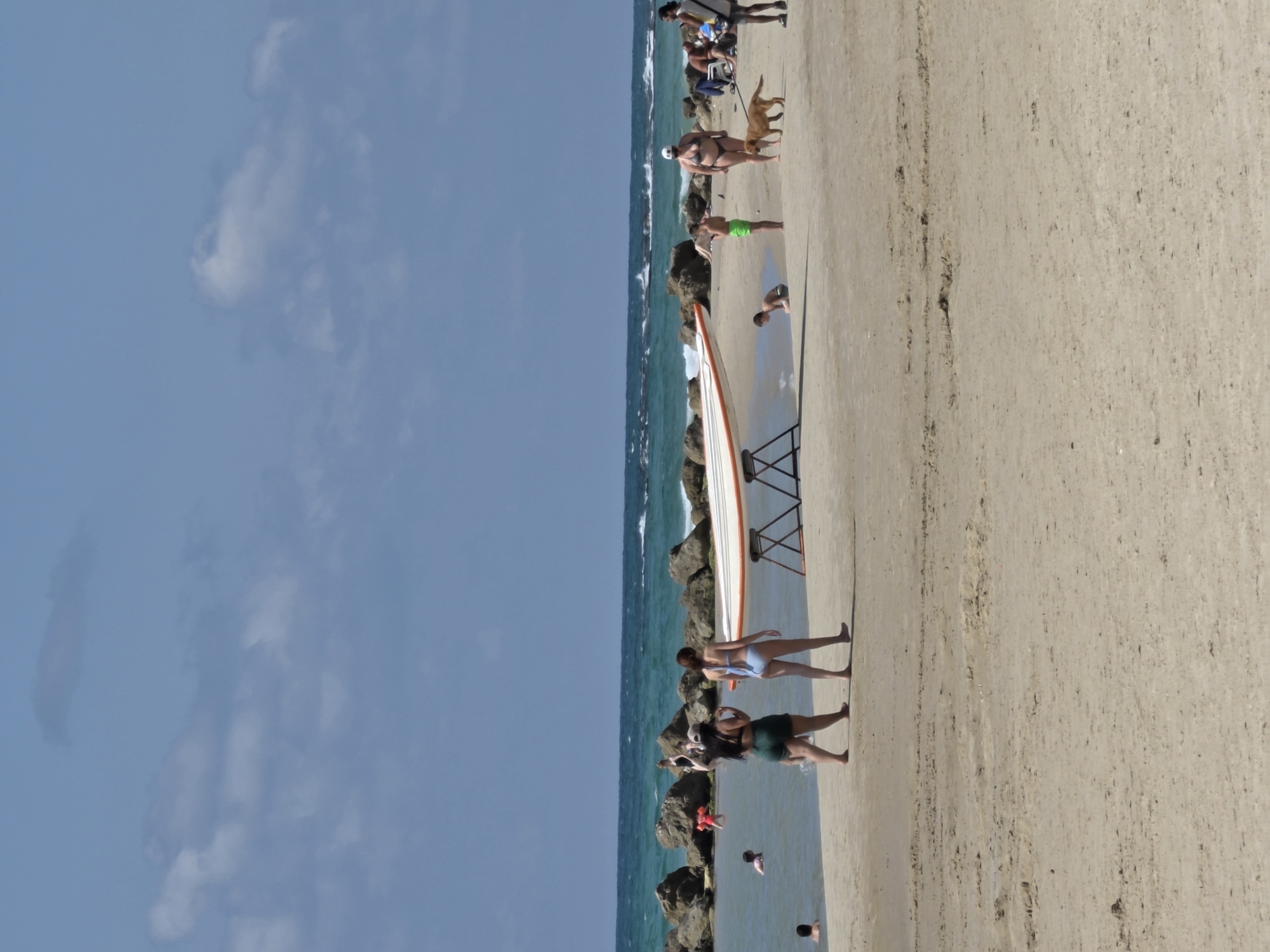 A photograph captures a sunny beach scene in Bat Yam, Israel. The sky is blue with scattered clouds to the left of the frame. The scene focuses on the beach, where a lifeguard boat rests on a stand. People populate the shoreline, some walking along the beach, others standing by the water's edge, and a dog is present. The beach runs along the right side of the image, and the ocean and rocks are to the left.
