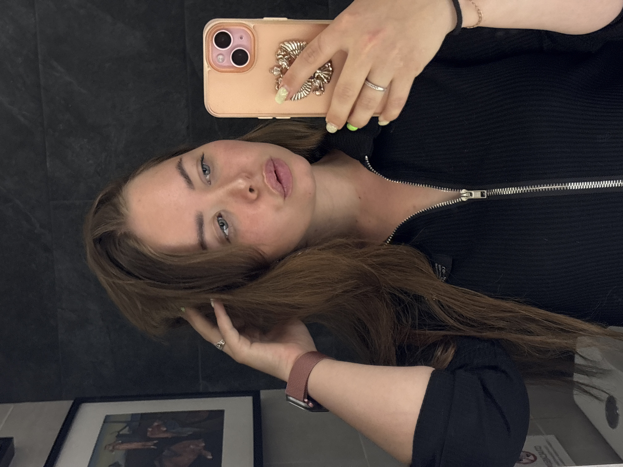 A young woman with long brown hair and blue eyes is taking a selfie in what appears to be a bathroom. She is holding a peach-colored iPhone 13 in her right hand, which has a decorative metallic design on the back. She is wearing a black zip-up jacket and a pink Apple Watch on her left wrist. She has rings on both hands. The background is a dark gray tiled wall. A framed picture is partially visible in the lower left corner. There is also a toilet and toilet paper dispenser visible on the bottom right. Based on the location data requested, the image was taken in the city of Chaikovsky, Russia.