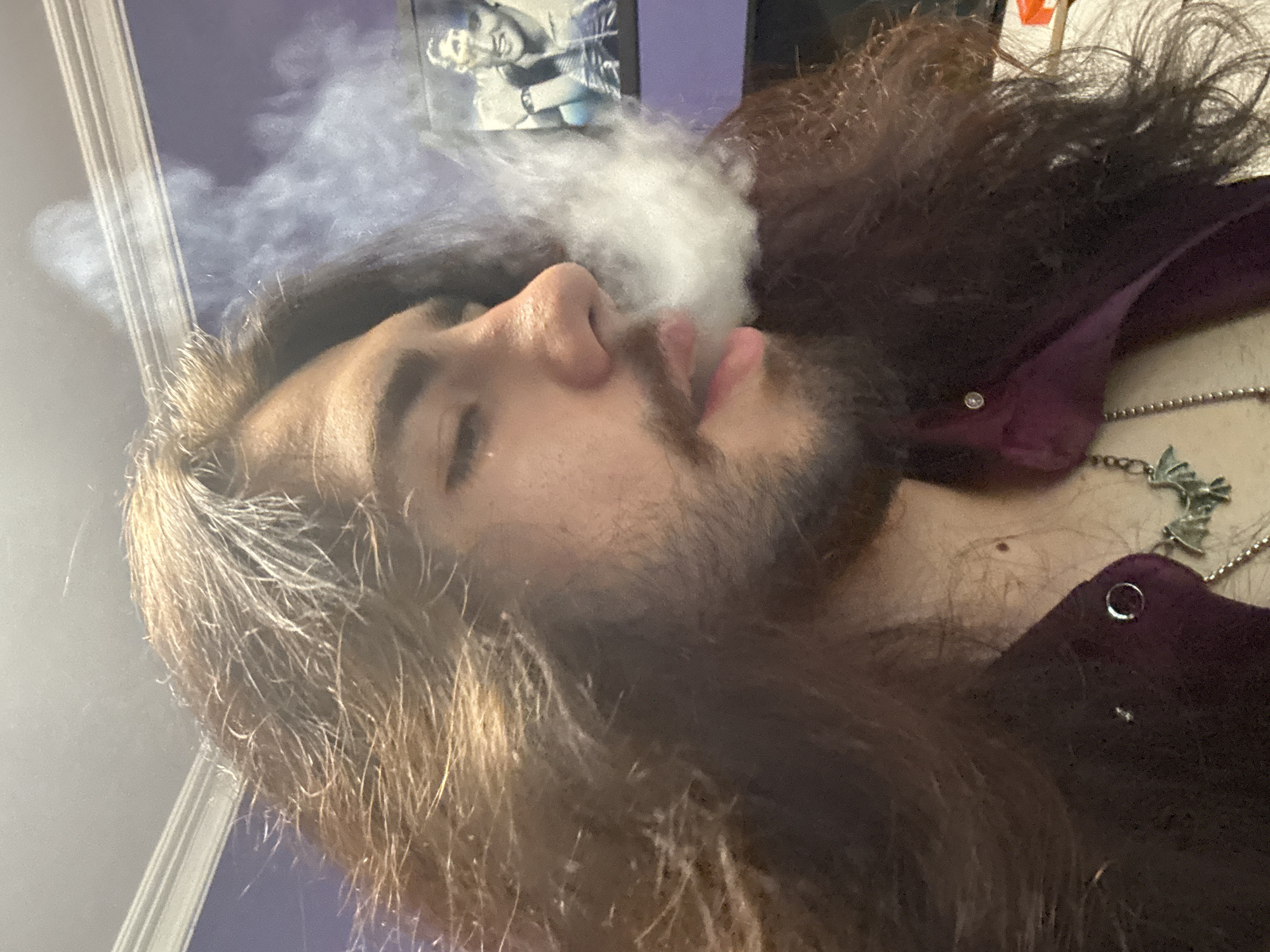 An individual with long, dark hair and a beard exhales a plume of smoke from their mouth. They are wearing a dark purple garment with a buttoned collar and a necklace featuring a bat pendant. In the background, a framed picture of a smiling man is visible on a purple wall. The lighting suggests an indoor setting, possibly a bedroom, with light emanating from above. The overall scene appears to be a candid portrait captured from a close-up perspective. The location is not discernible from the image.