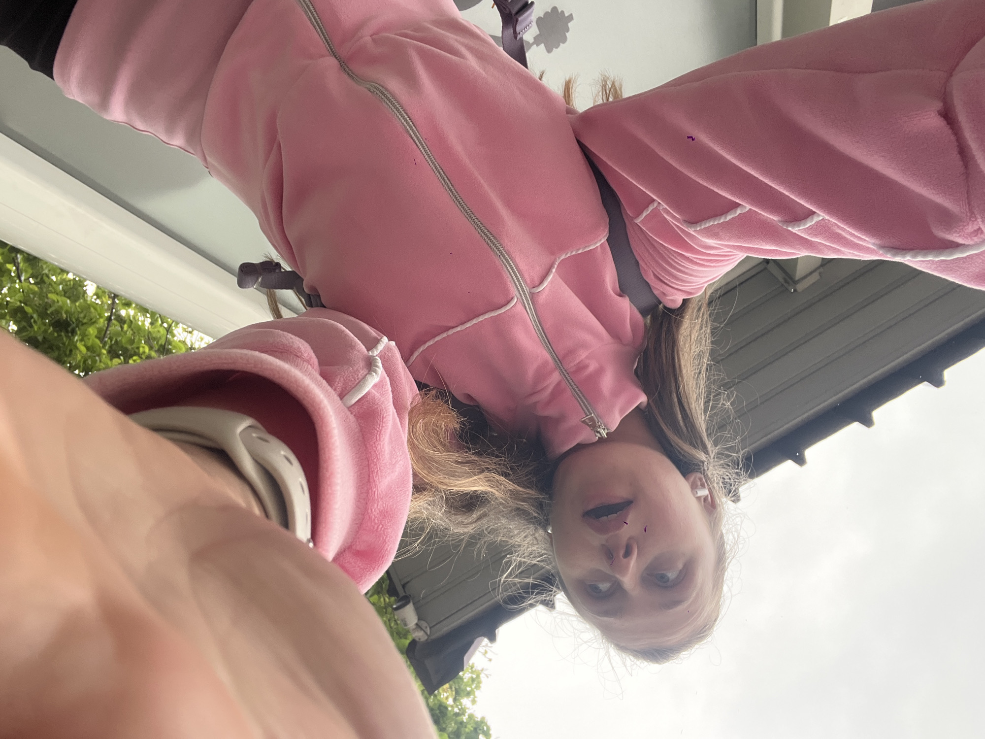 A young person with blonde hair is looking down at the camera. They are wearing a pink zip-up jacket with white drawstring. A white smartwatch is visible on their left wrist. The background includes a white exterior wall and green foliage suggesting an outdoor setting. The location is Bjeliši, Montenegro.