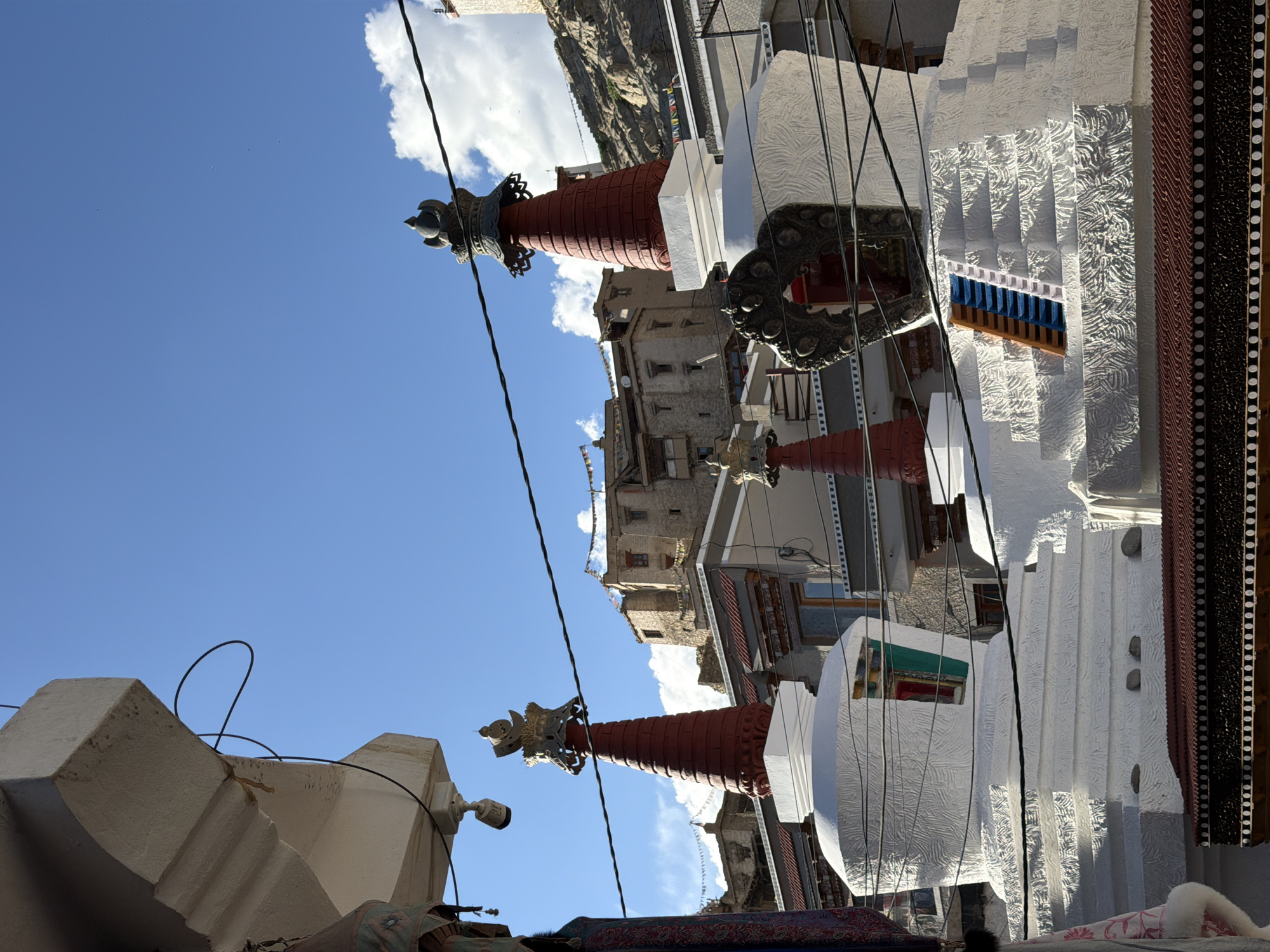 The image shows a view of the Leh Palace in Leh, India. The palace is a tall, multi-story structure with a fortress-like appearance, built on a hillside. In the foreground, several stupas, which are Buddhist reliquary monuments, are visible. These stupas are primarily white with red spires and ornate golden finials. Some of the stupas have intricate patterns on their surfaces. A ladder with blue and orange steps is placed on one of the stupas. The sky above is a clear blue with a few white clouds. Wires are strung across the scene, suggesting the presence of electricity. The overall scene depicts a cultural and architectural landscape typical of Leh.