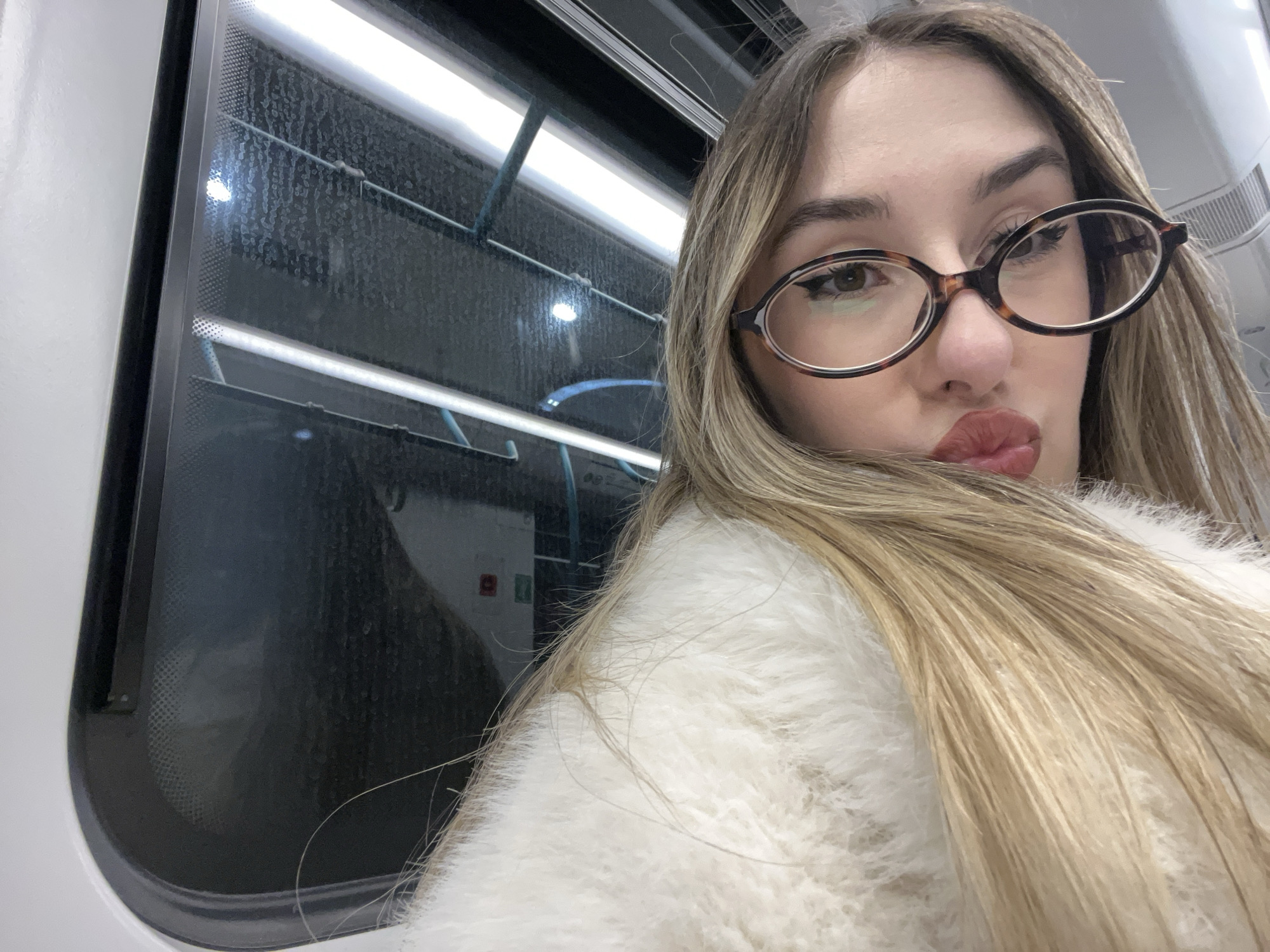 A close-up selfie captures a young woman inside what appears to be a public transport vehicle in Sofia, Bulgaria. She has long, straight, light brown hair and wears distinctive round, tortoise-shell patterned glasses. Her eyes are light brown, accented with eyeliner, and her lips are full, made up with reddish-brown lipstick, forming a pouty expression directed at the camera. She is dressed in a fluffy, white or cream-colored garment.
The background is dominated by a large window of the vehicle, which appears streaked and dirty, possibly from rain or moisture on the exterior. Bright, linear artificial lights illuminate the interior from above and below a horizontal bar visible through the window. These lights reflect on the window's surface, which also displays a dot-patterned frit. Below the window, a dark panel features two small, square indicators, one red and one green, suggesting some form of control or emergency system within the vehicle. The view outside the window is dark and indistinct, indicating it might be night, inside a tunnel, or in an environment lacking natural light. The setting, with its internal lighting and window structure, strongly suggests a metro, train, or tram carriage. No visible text can be clearly discerned on any objects in the image.