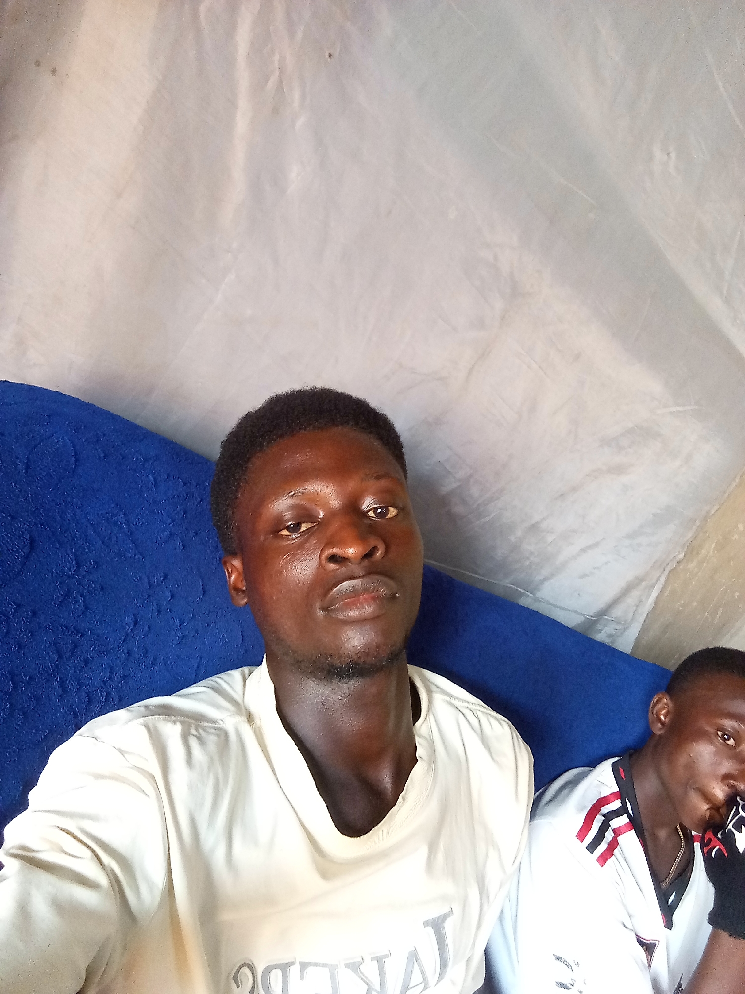 Two young Black males are seated side-by-side indoors, with a bright blue cushioned surface behind them and a wrinkled off-white fabric backdrop. The male on the left is in the foreground, facing the camera. He has dark skin, short black hair, and is wearing a light-colored t-shirt with faded gray text on it. The male on the right is partially visible in the background, looking towards the left, and is wearing a white shirt with red and black accents on the collar and a black sleeve. The environment appears to be a simple, possibly makeshift, indoor space.