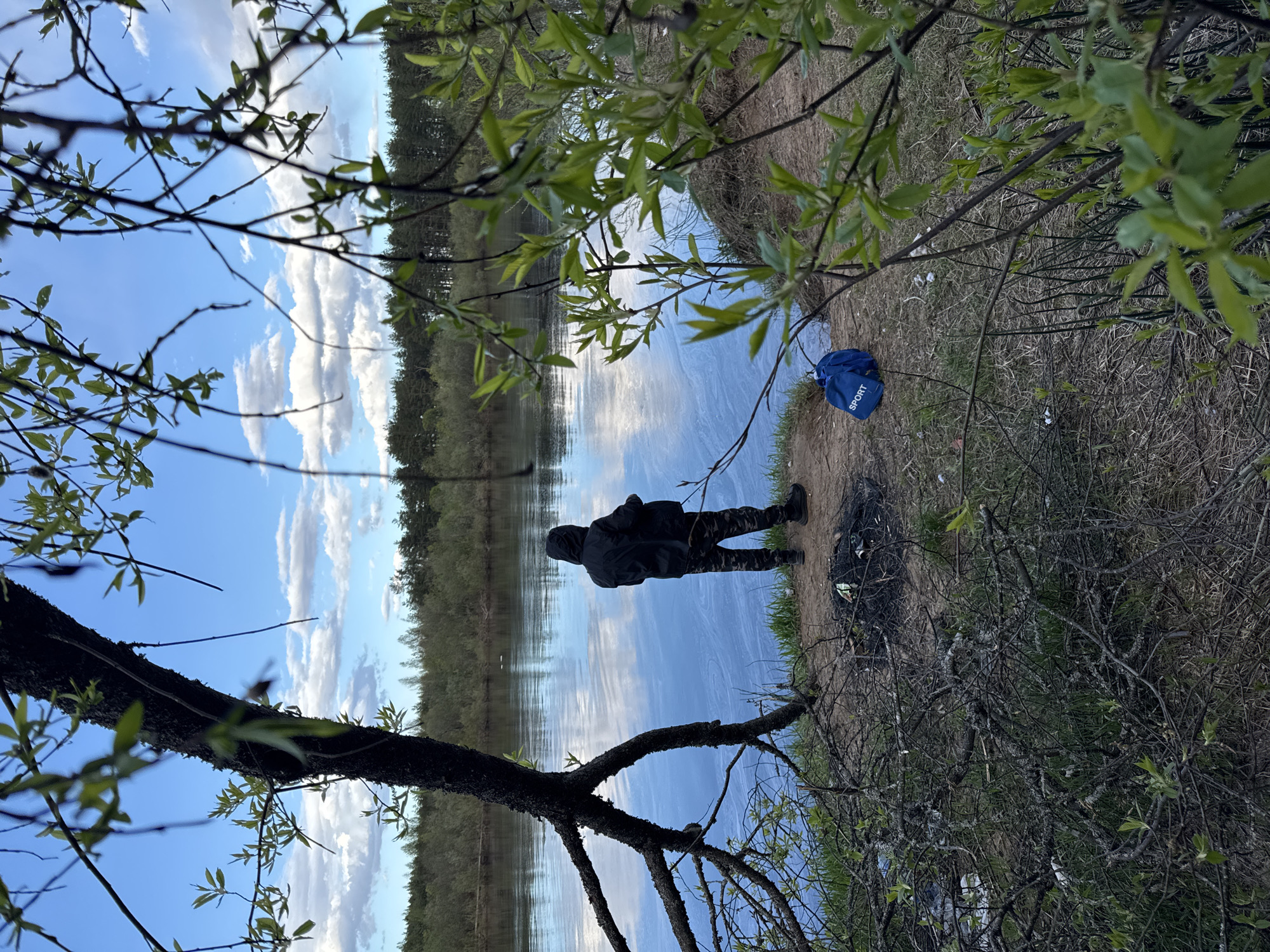 An outdoor scene, possibly in the vicinity of Малая Рукавицкая, Russia, depicts a body of water reflecting a partly cloudy sky. A person stands on the bank, silhouetted in dark clothing. A blue "SPORT" bag is visible near the edge. The scene is framed by branches and foliage.