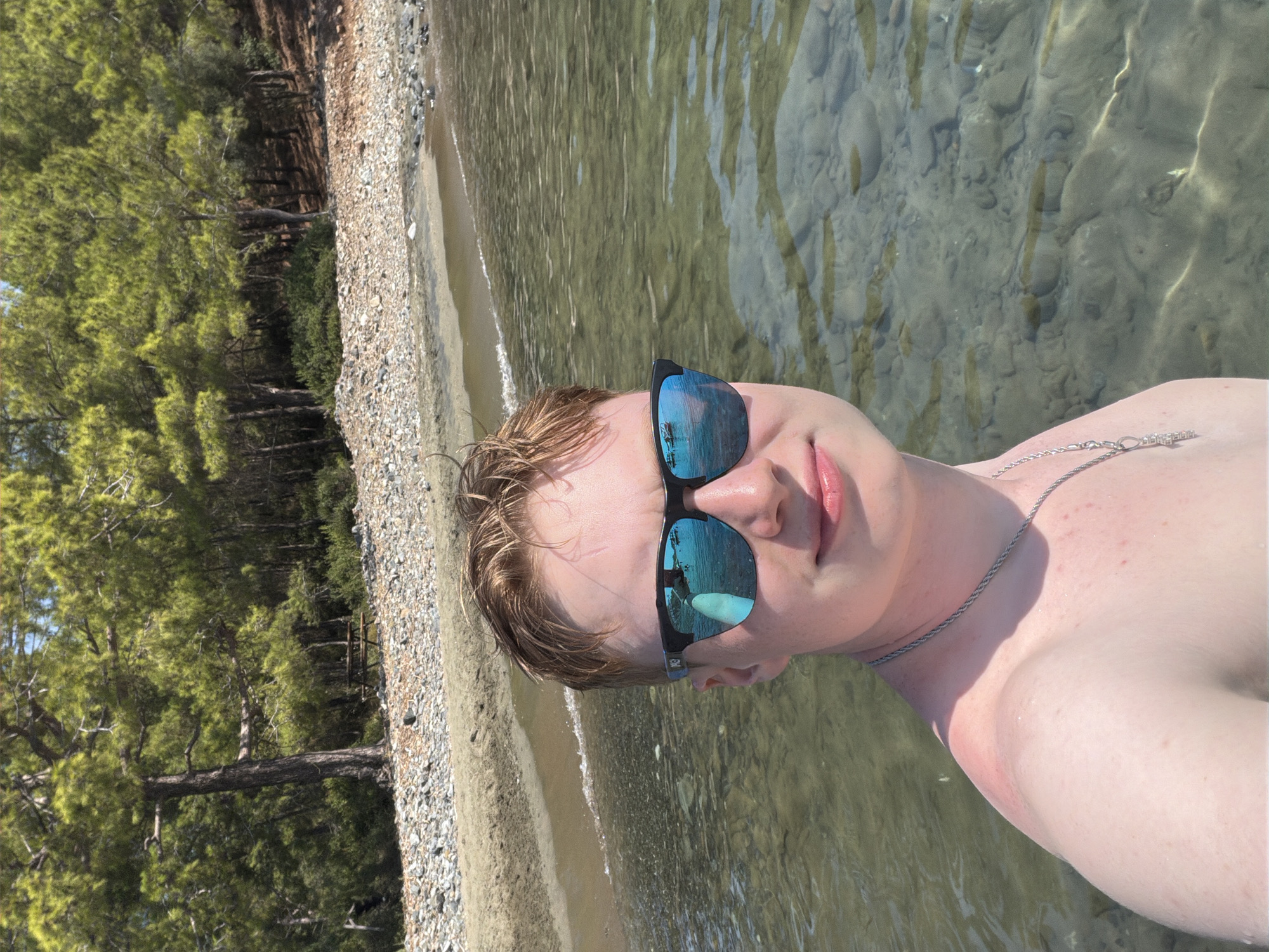 This is a selfie taken from a low angle, with the subject partially submerged in clear, shallow water. The person is wearing sunglasses with blue reflective lenses, and a silver chain necklace. They have light-colored hair and fair skin, with a light tan on their shoulder. The background shows a pebbly beach and a dense green forest with tall trees, characteristic of a Mediterranean coastal environment like Kemer, Turkey. The water is calm and reflects the sunlight, illuminating the rocky seabed below. The overall impression is one of a relaxed summer day at the beach, likely during daylight hours given the bright lighting. The subject's expression is neutral, and the scene evokes a sense of peace and enjoyment of nature. No text is visible in the image.