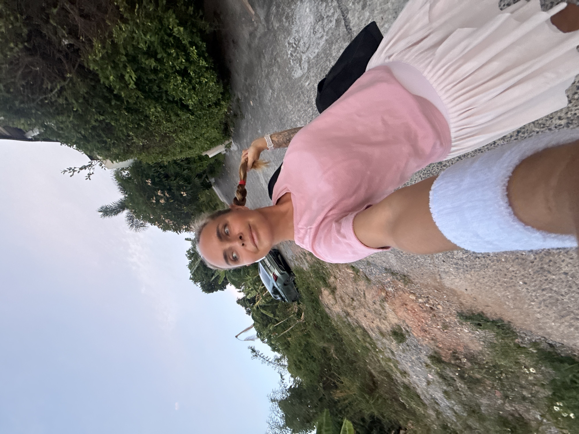 In the city of Ko Samui District, Thailand, a woman is captured in a selfie on a gravelly, uphill road. She is wearing a pink t-shirt and a pale pink pleated skirt. Her hair is pulled back into a braid with a red elastic. A black bag is visible behind her. In the background, lush green foliage lines the road, and a dark-colored car is parked on the incline. The sky above is a pale blue with some wispy clouds. The overall lighting suggests it is either early morning or late afternoon.