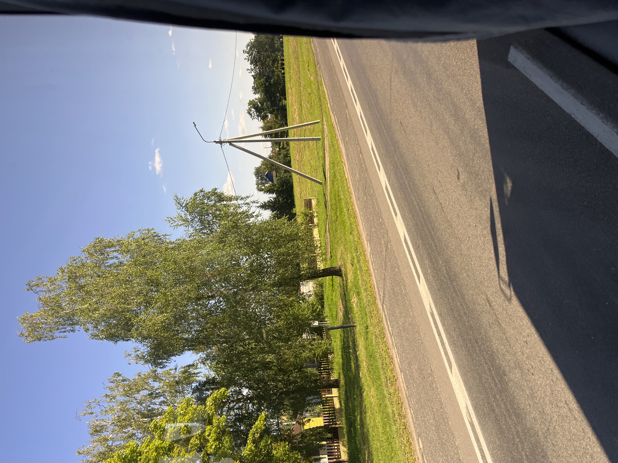 The image depicts a sunlit day in Myadzyel, Belarus, with a view from a vehicle traveling along a paved road. The road has white dashed lines marking the lanes. To the left of the road, there is a grassy area with a fence and a yellow house. Several trees, including birches, are present. Further into the background, there is a field with a triangular utility pole holding a street light and power lines. The sky is blue with a few scattered white clouds. The right side of the image is partially obscured by the dark interior of the vehicle.