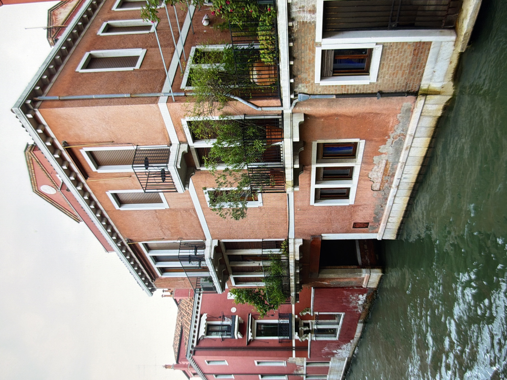 The image shows a canal with buildings on either side. The buildings are made of brick and have a reddish-orange hue. There are windows and balconies on the buildings, and some of the balconies have plants on them. The canal is a dark green color, and there are ripples on the surface of the water. The sky is overcast and a light gray color. The scene appears to be in Venice, Italy, not Kyiv, Ukraine.