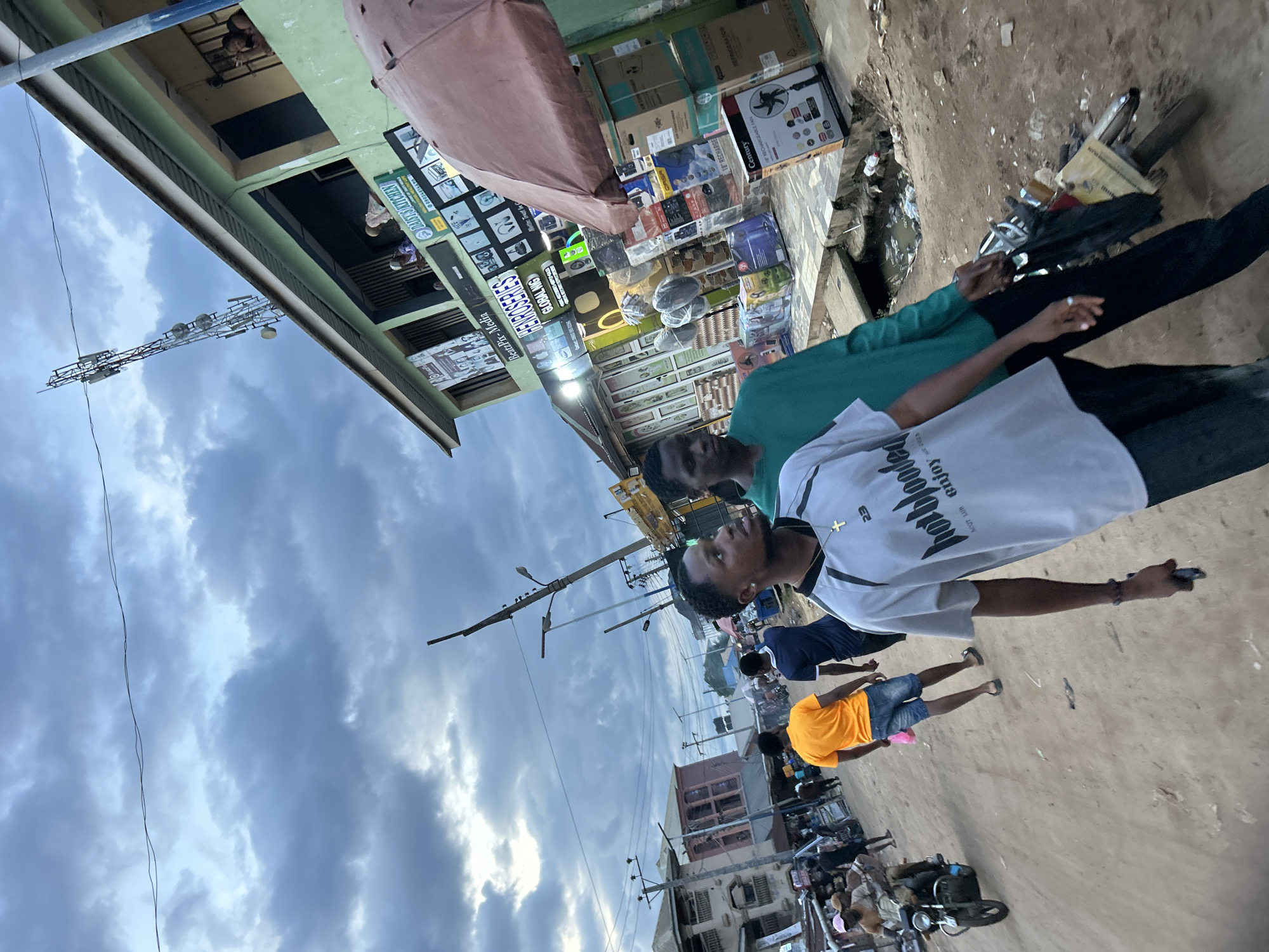The image, taken in Ihiagwa, Nigeria, captures a bustling street scene, likely a commercial or market area, under an overcast sky. The ground appears unpaved and dusty, with visible debris and an open drainage ditch running alongside the buildings.
In the foreground, a young man wearing a white t-shirt with "hot blooded ACQT LUB enjoy" printed on it, a cross symbol, and "23", is prominently featured. He is looking forward, with his arm extended slightly. Beside him, another man in a green long-sleeved shirt is also visible, partially obscured. Other people are scattered further down the street, including one person in an orange t-shirt and blue shorts. Motorcycles are parked or being ridden along the street.
The street is lined with buildings and makeshift stalls displaying various goods. Visible items include numerous boxes, likely containing electronics or household appliances, such as a "Century RECHARGEABLE FAN" and a "REFRIGERATOR". Signs on the shops and stalls show text like "BeatzPix-Media," "HENROSERIES," "GLOBAL NIG," and "DAISY'S KITCHEN". One sign partially reads "Motto: Trust In".
In the background, a tall communication tower or electricity pylon stands against the cloudy sky. The lighting suggests an overcast day, possibly late afternoon or early morning, with soft, diffused light. The overall scene depicts typical daily activity in a lively urban or semi-urban setting.