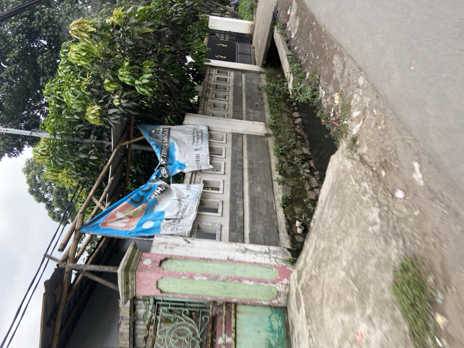 The image shows a street in Margaasih, Indonesia (latitude: -6.962, longitude: 107.548). A concrete drainage ditch runs parallel to the street, with a concrete sidewalk to the right. The ditch is filled with murky water and contains some trash. A large tree with green leaves grows on the left side of the image, casting shadows on the side of the road and the drainage ditch. A concrete wall with a gate in the middle separates the road from a property on the left. The gate appears to be closed. Two large blue and white signs, presumably for a local business, are attached to a wooden frame above the wall. There are no visible people in the image. The sky is overcast. The overall scene is typical of a residential street in a developing country.