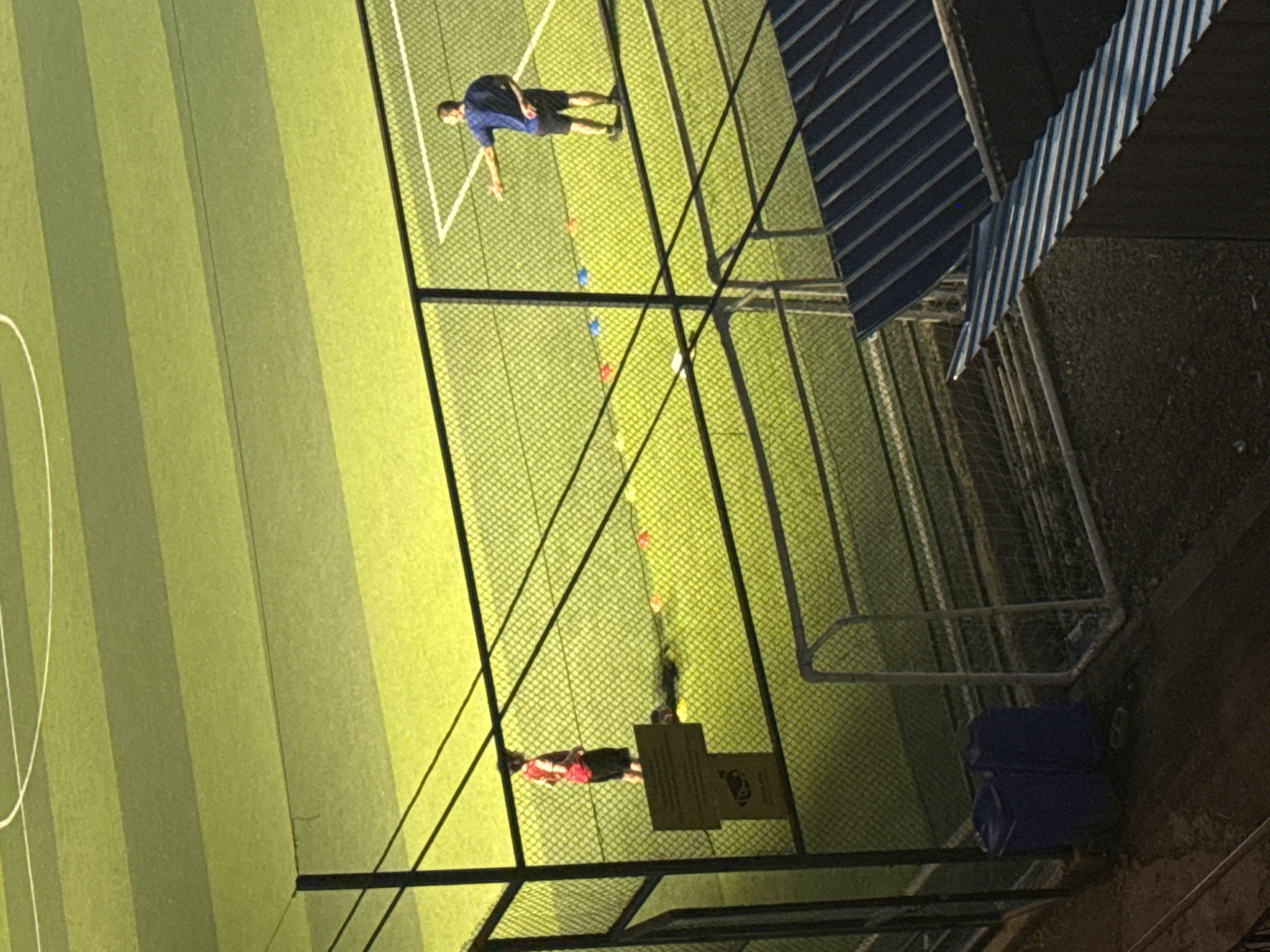 The image shows an outdoor artificial turf soccer field at dusk, likely in Tbilisi, Georgia, given the presence of distinctive blue recycling bins often seen in the city and the general urban environment. The field is sectioned by white lines and appears to be fenced off.
In the foreground, two large blue cylindrical recycling bins are positioned on a dark, uneven surface that appears to be dirt or gravel. To the right of the bins, a portion of a soccer goal with a white frame and net is visible. A metal roof structure with corrugated blue and silver panels partially obscures the view of the upper part of the field.
On the field itself, behind a chain-link fence, two individuals are present. On the left, a young person wearing a red and black soccer uniform stands near a yellow sign with an image of a security camera. To the right, a man in a dark blue shirt and black shorts stands facing away from the camera, with his right arm extended, possibly directing activity. Several brightly colored training cones (red, blue, and orange) are scattered on the turf between the two individuals. Shadows are cast on the field, indicating the time of day. The overall lighting suggests late afternoon or early evening.