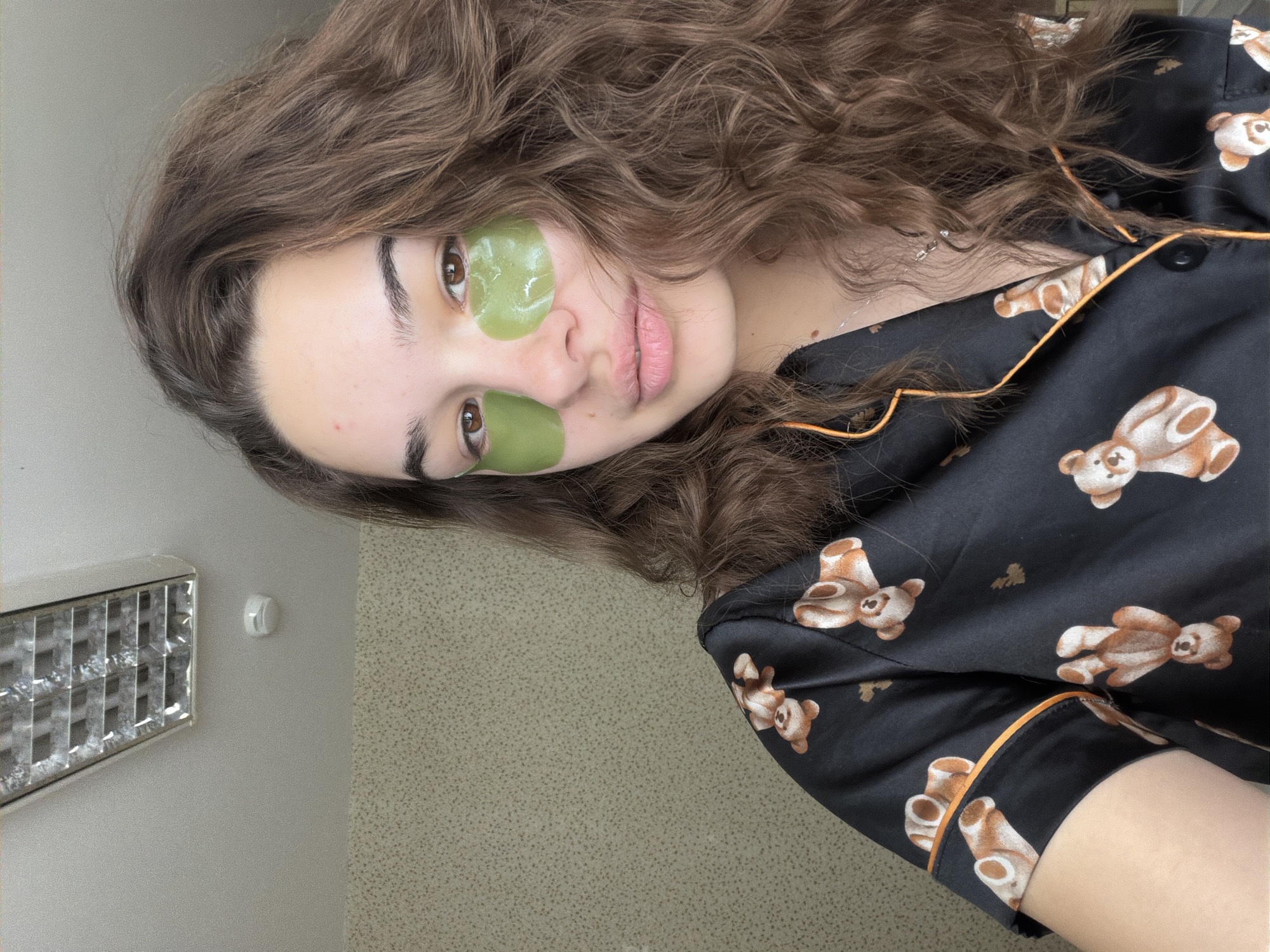 A selfie of a young woman. She has light skin, brown eyes and curly brown hair. She is wearing green eye masks under her eyes. She wears a black pajama top with a teddy bear print and orange piping. The background is a light beige wall with a light fixture. The location is not discernible.