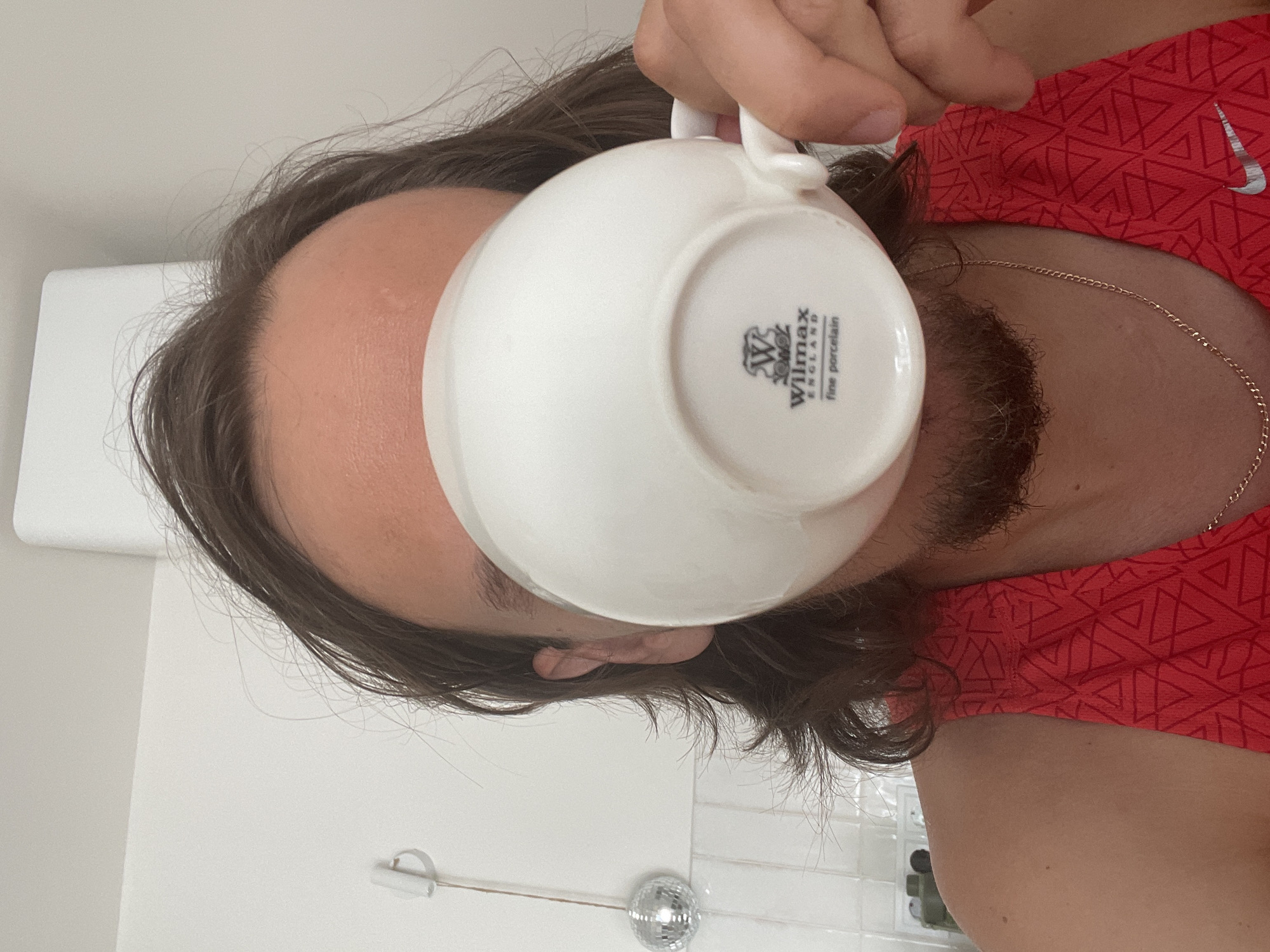A person is holding a white "Wilmax" fine porcelain teacup to their face, obscuring their mouth and nose. The person has brown hair and a beard. They are wearing a red shirt with a pattern and a gold chain necklace. The background shows a white wall, a light fixture, a disco ball, and some bathroom items. The image doesn't provide any information to verify a location in Izhevsk, Russia.