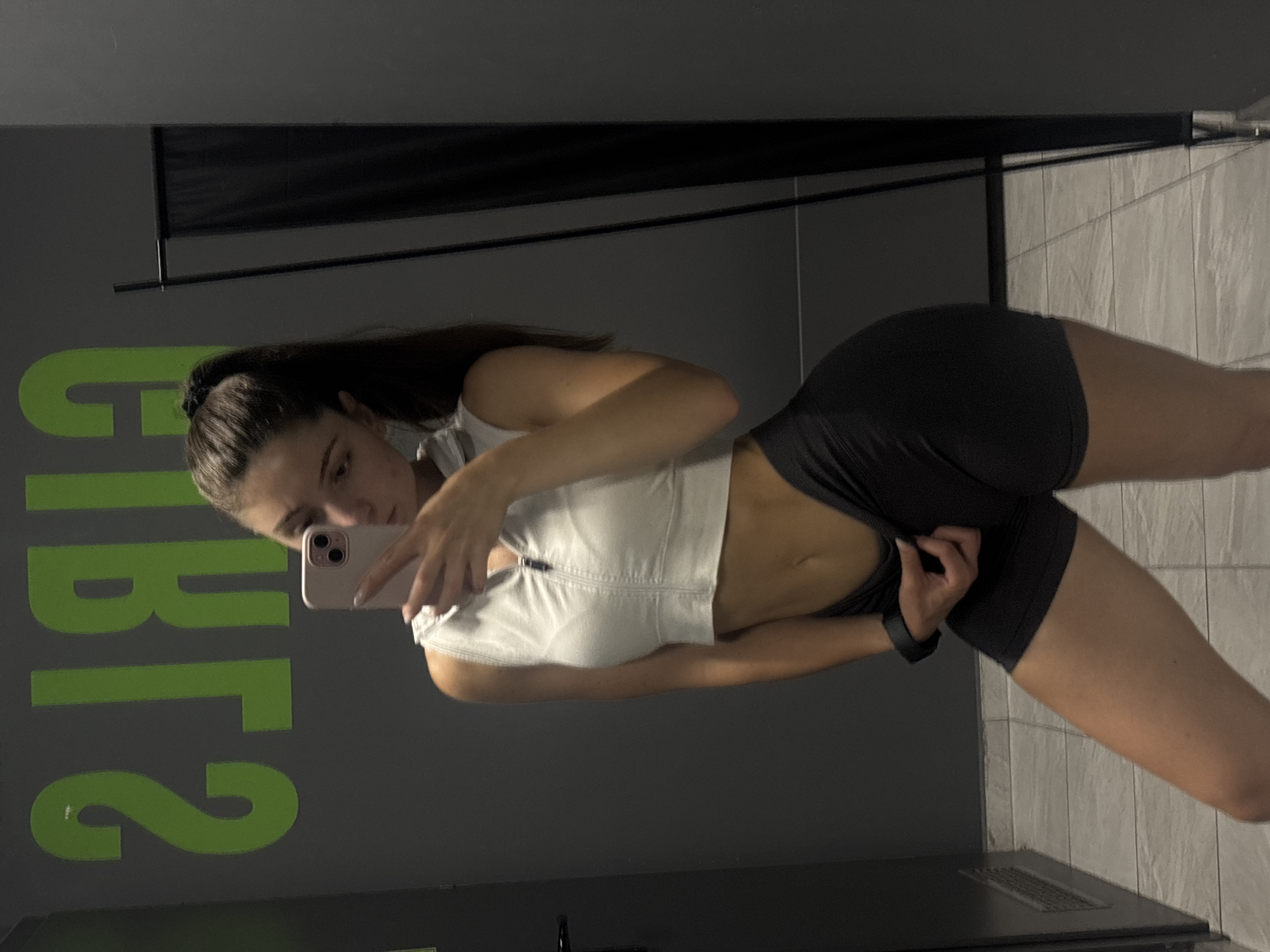 In a gym changing room with grey tiled flooring and dark grey walls, a young Caucasian female with brown hair pulled into a ponytail, wearing a white cropped zip-up athletic top and black athletic shorts, is taking a selfie in a mirror. Green lettering is visible on the wall behind her, which appears to be reversed in the mirror's reflection. The overall lighting is dim. This scene is likely from the "Gibs" gym located in Varna, Bulgaria, based on the visible lettering and common gym aesthetics.