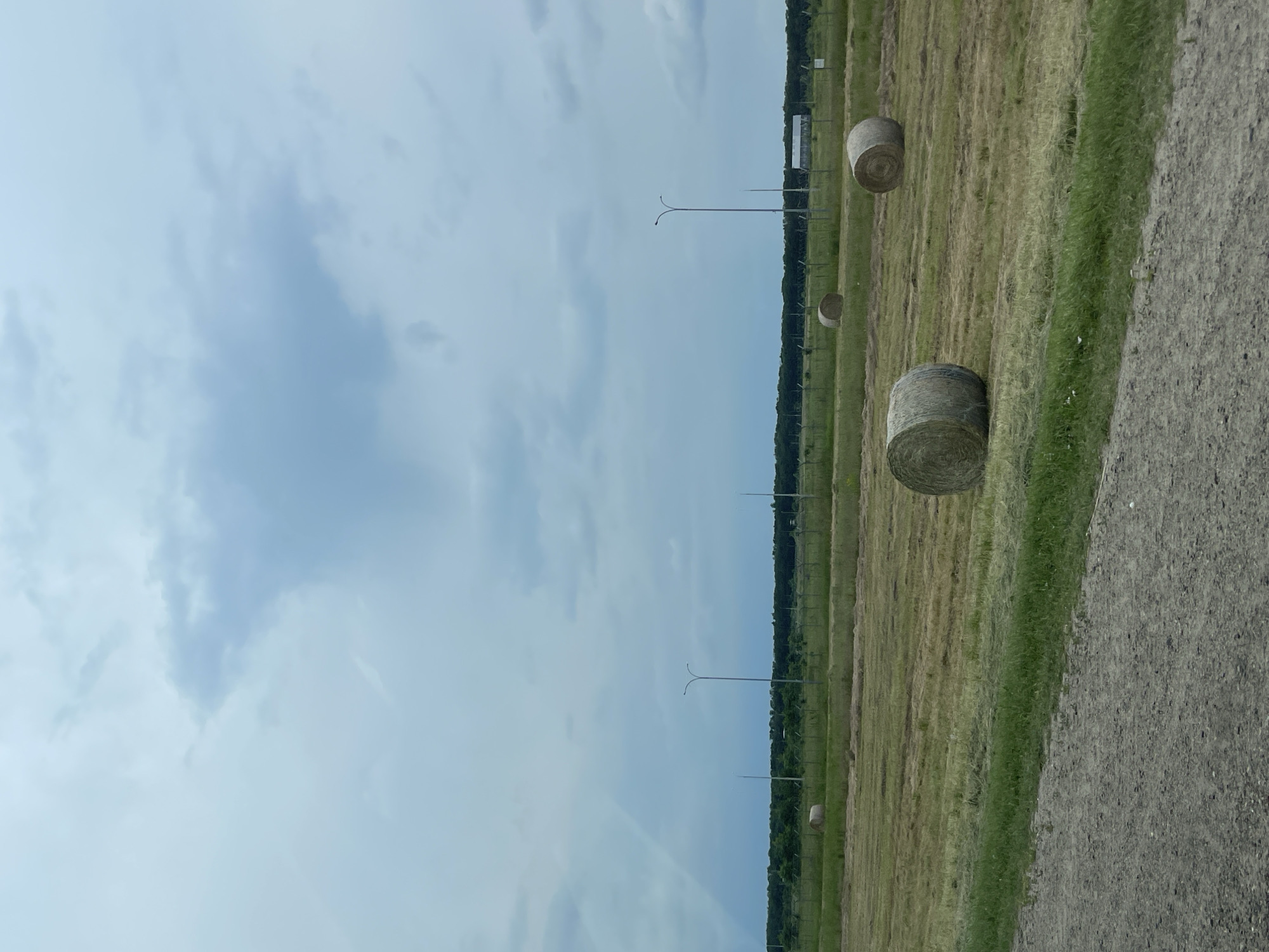 An outdoor shot captures a rural scene under a cloudy sky. The foreground features a gravel road bordering a field of dry, cut grass. Several round bales of hay are scattered across the field. In the middle ground, a fence line with tall poles and barbed wire separates the field from a treeline in the background. A building is visible behind the fence line. The sky is a mix of grey and blue clouds, with a hint of a rainbow on the left side. The overall impression is that of an agricultural landscape in a somewhat overcast environment. The location is the city of Mikhaylovsk, Russia.