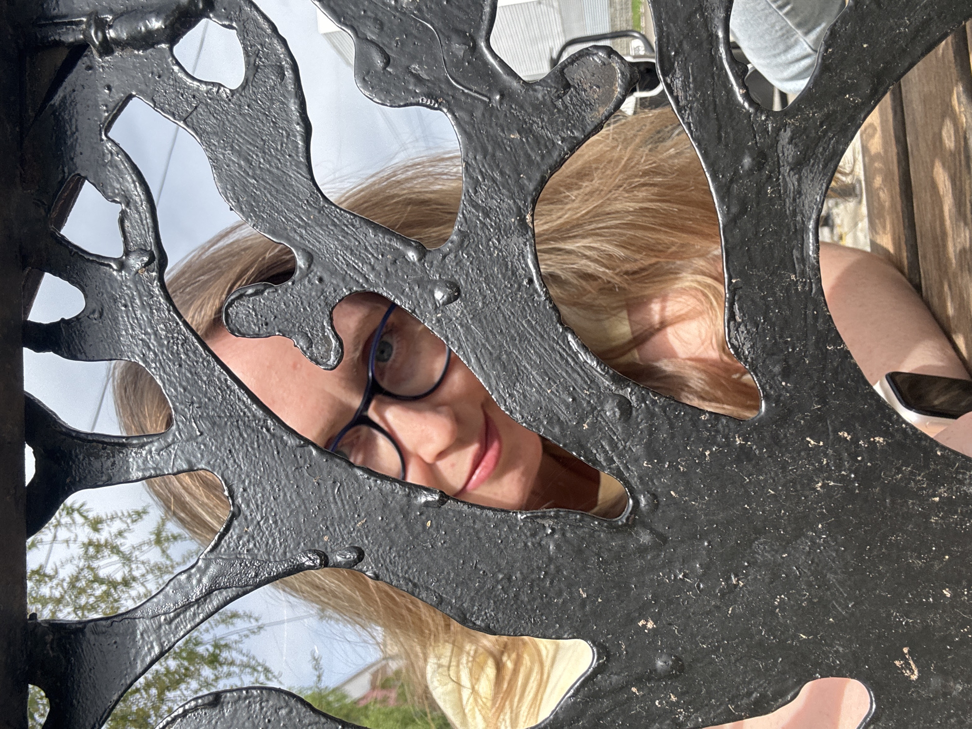 A person with blonde hair and glasses is peering through the ornate metalwork of an object. The metalwork is black and appears to be patterned like the branches of a tree. Outside the metalwork, elements of the environment are visible, including sky, trees, and what seems to be a building. The scene is shot from a low angle, looking up at the subject through the metalwork. The location is indicated as the city of Chaikovsky, Russia.